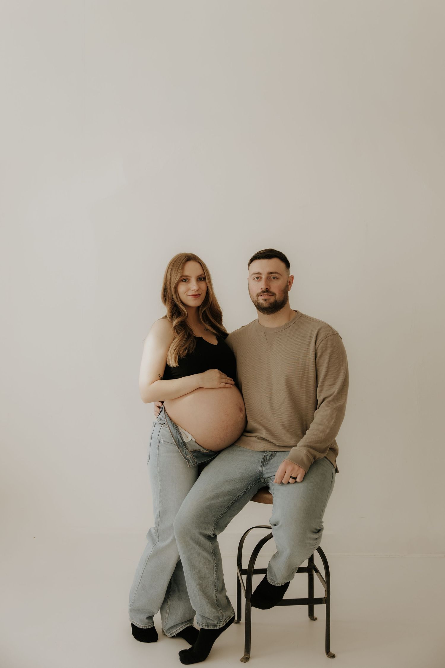 Maternity studio photo with couple sitting on black stool against white background.