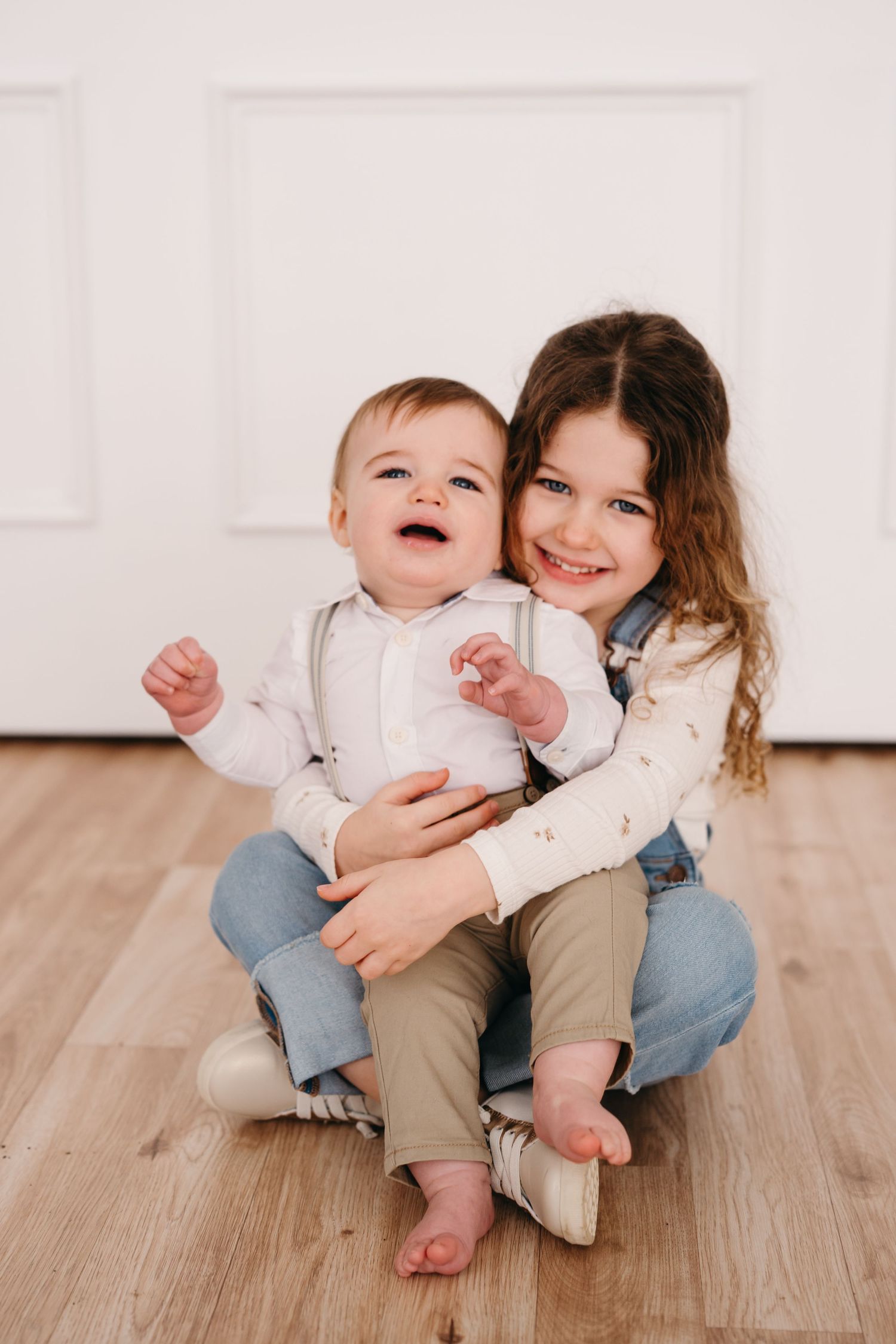 Two children sit together on a hardwood floor against a white wall, sharing a joyful moment.