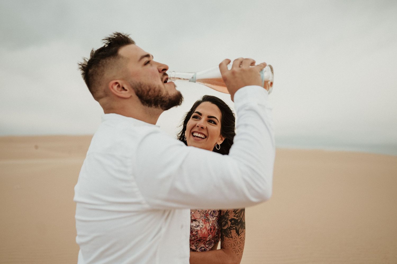 A couple shares a lighthearted moment drinking from bottles on desert sand dunes.