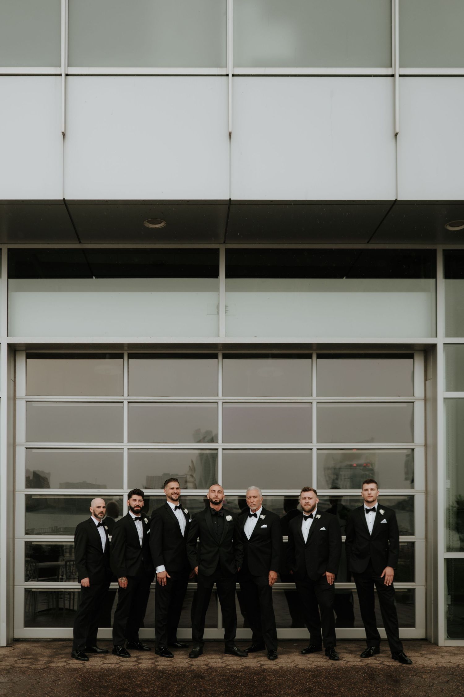 Groomsmen in classic black tuxedos pose in front of modern architectural building.