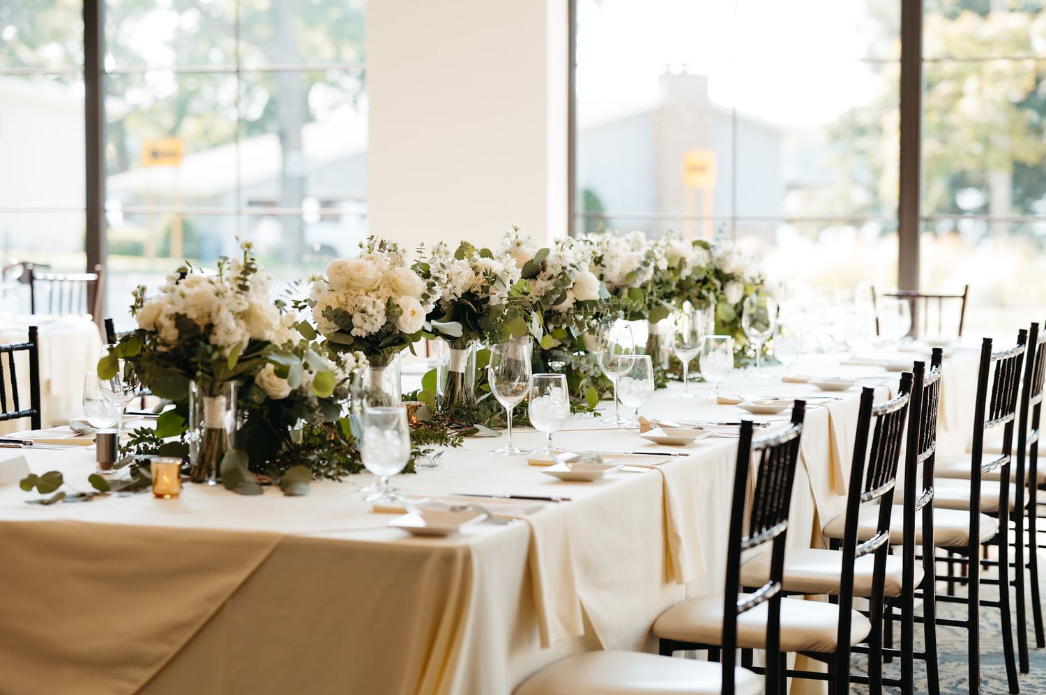 Elegant wedding reception table set with white linens and lush floral centerpieces near bright windows.