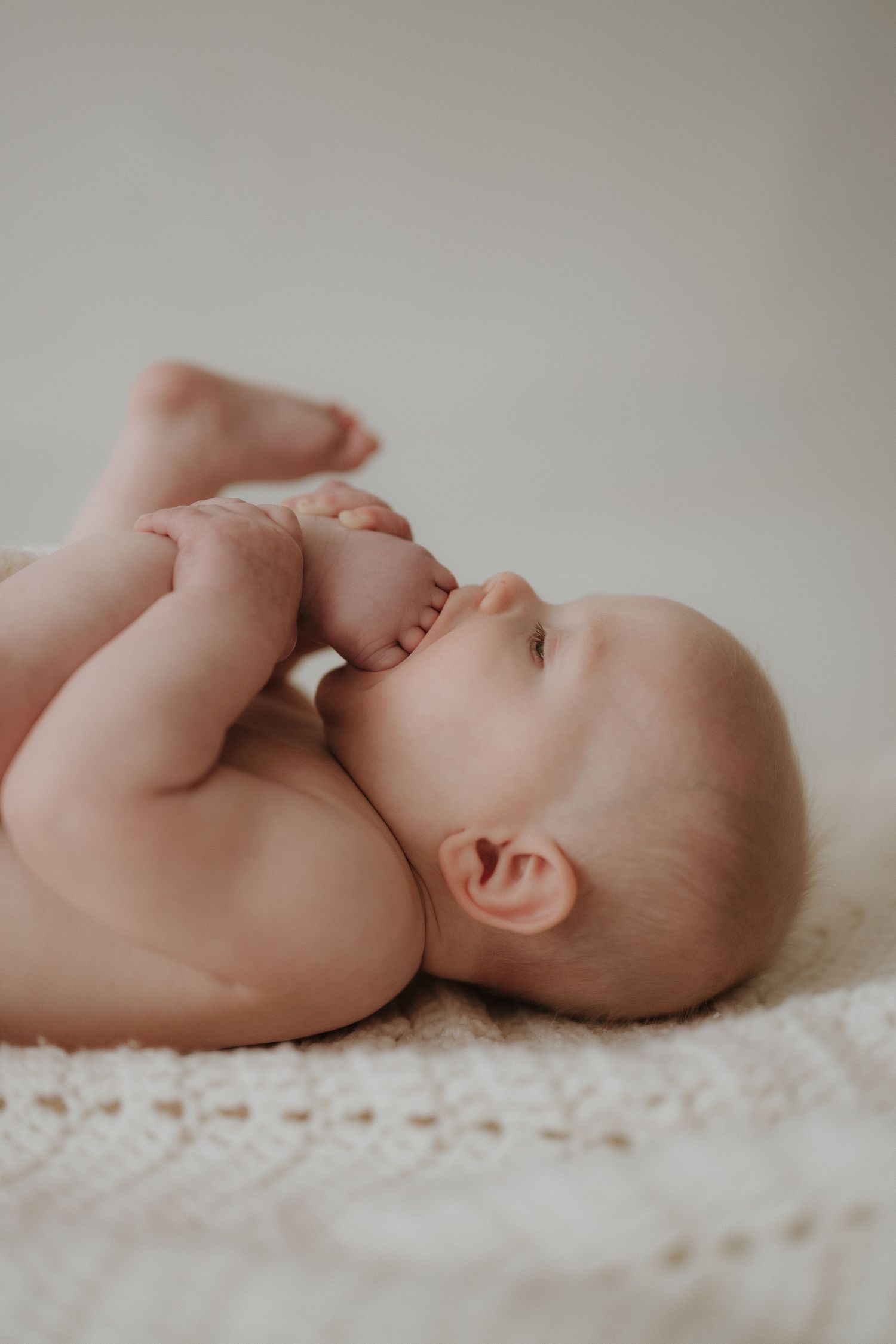 Naked baby lying on white knit blanket, holding feet up to mouth.