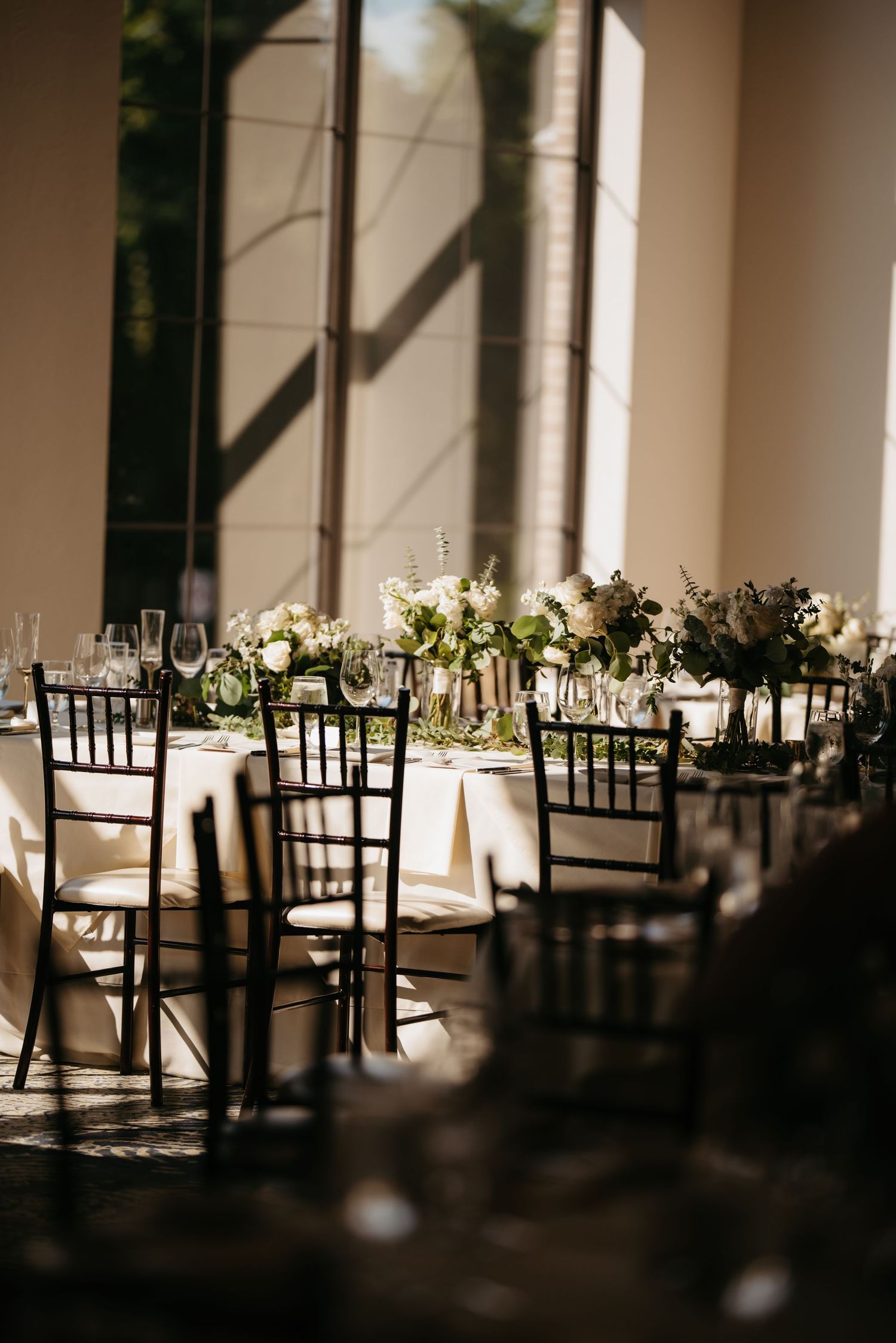 Elegant wedding reception tables set with black chairs and white floral centerpieces in a sunlit venue.