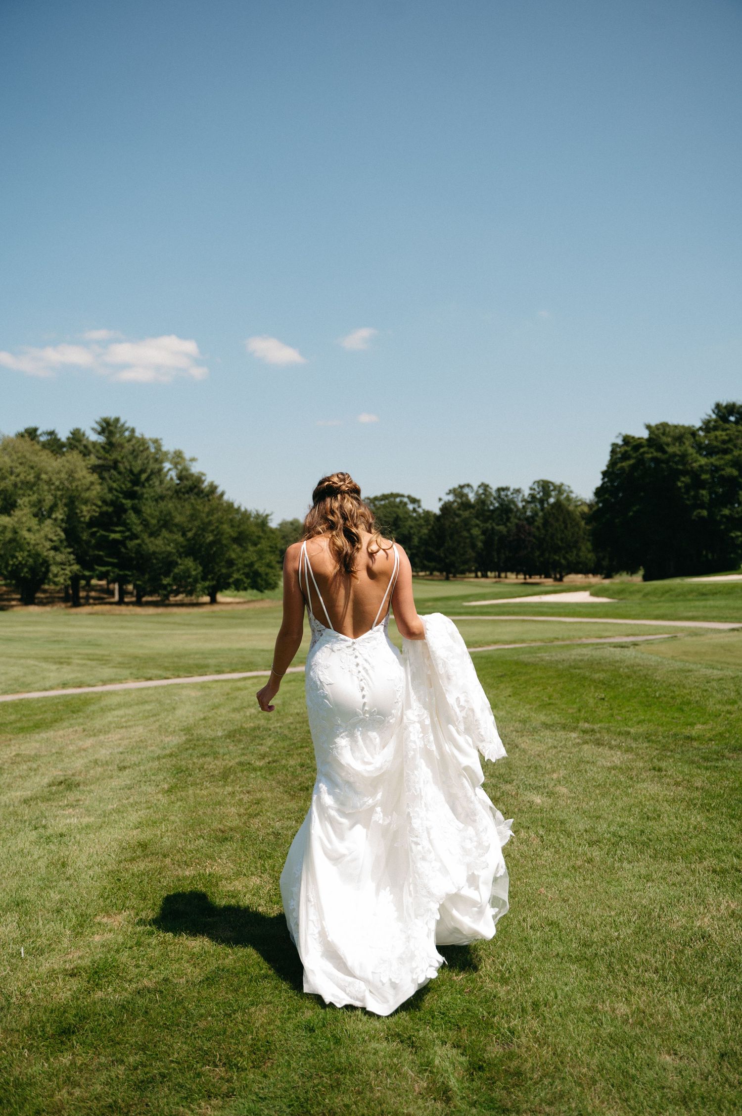 Two people in white dresses walking hand in hand across a sunny golf course with blue skies overhead.