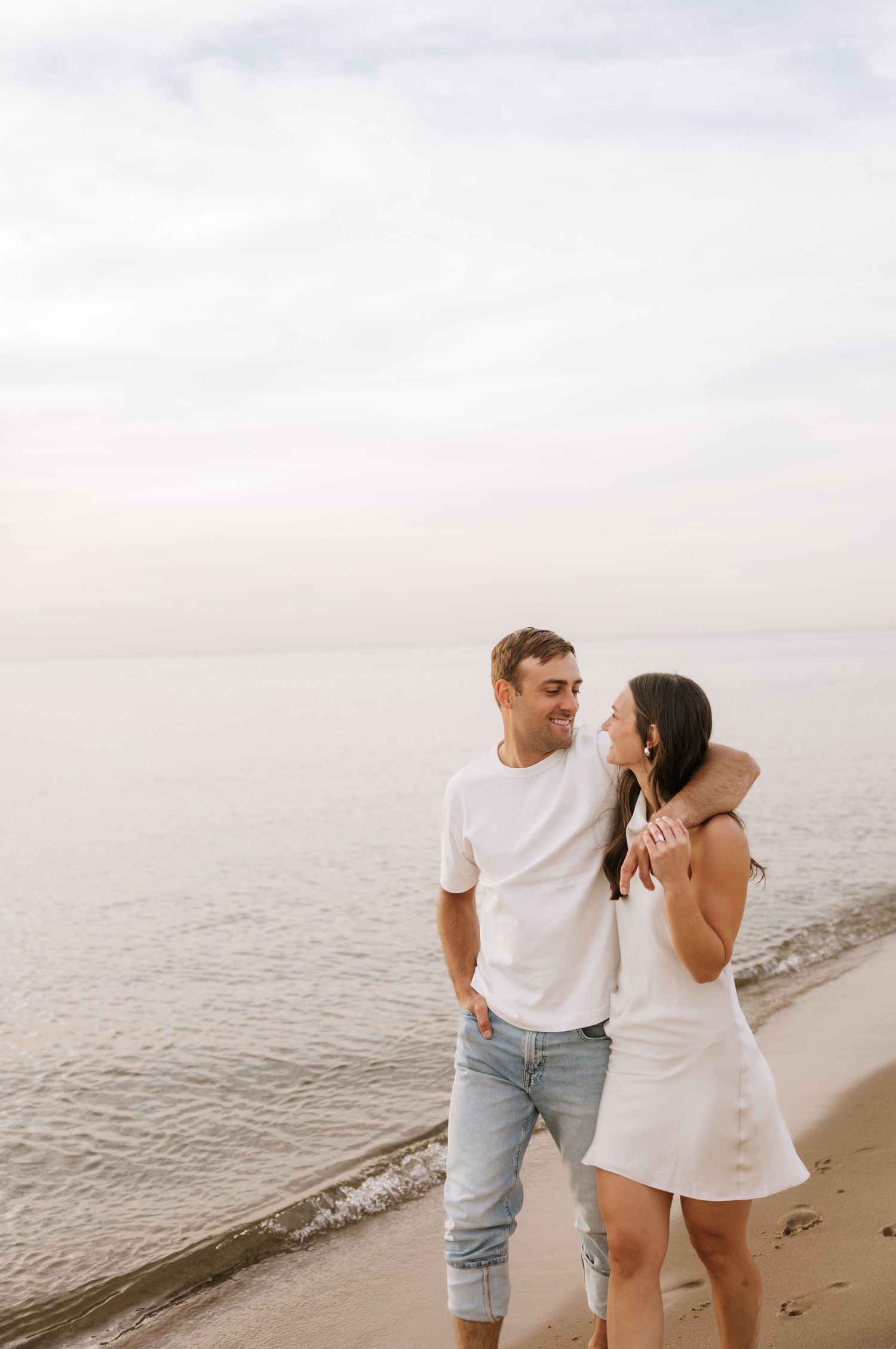 Romantic couple standing close together on a beach shoreline, gazing at each other in soft evening light.