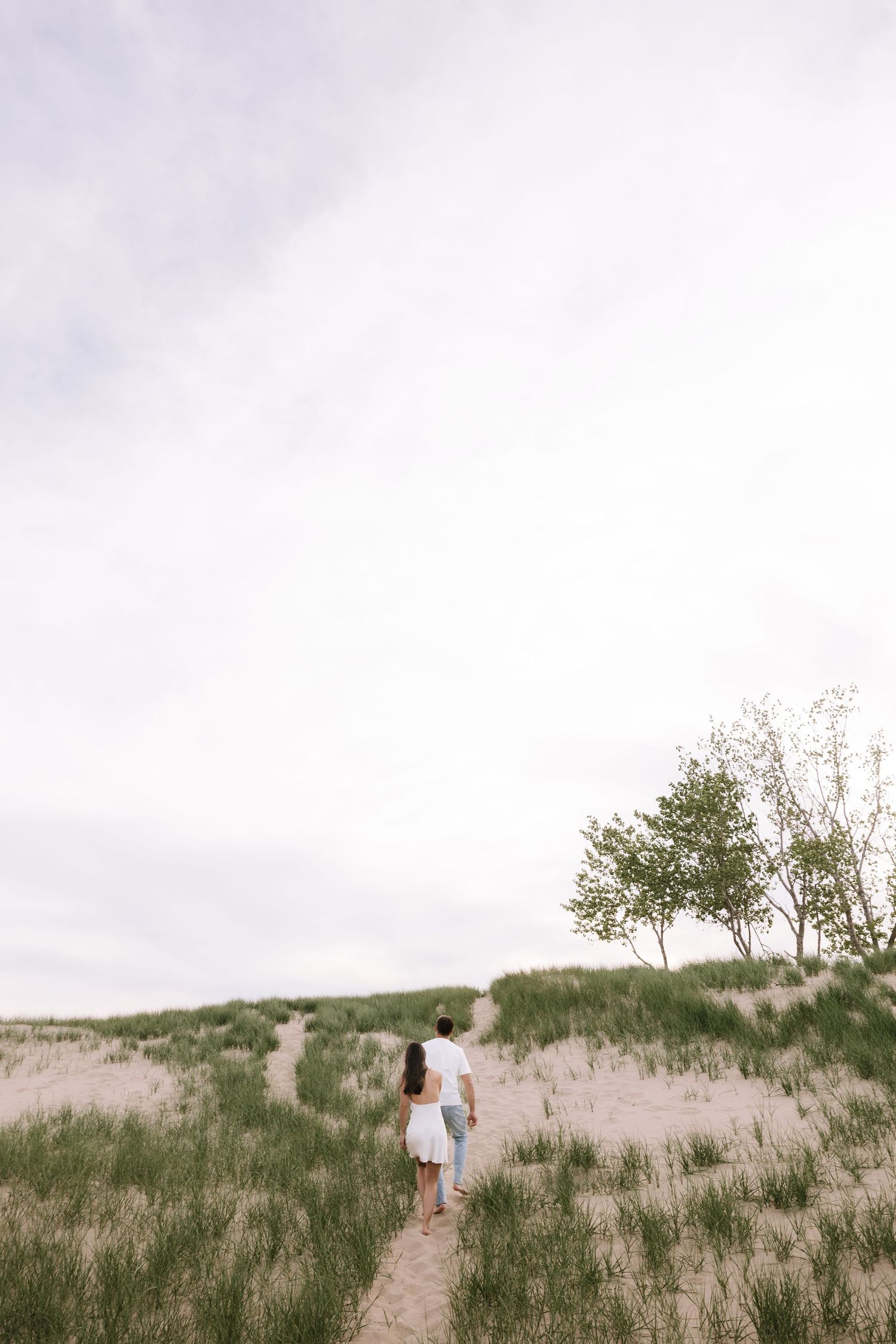 Couple walking hand in hand along a sandy path through green dunes under a cloudy sky.