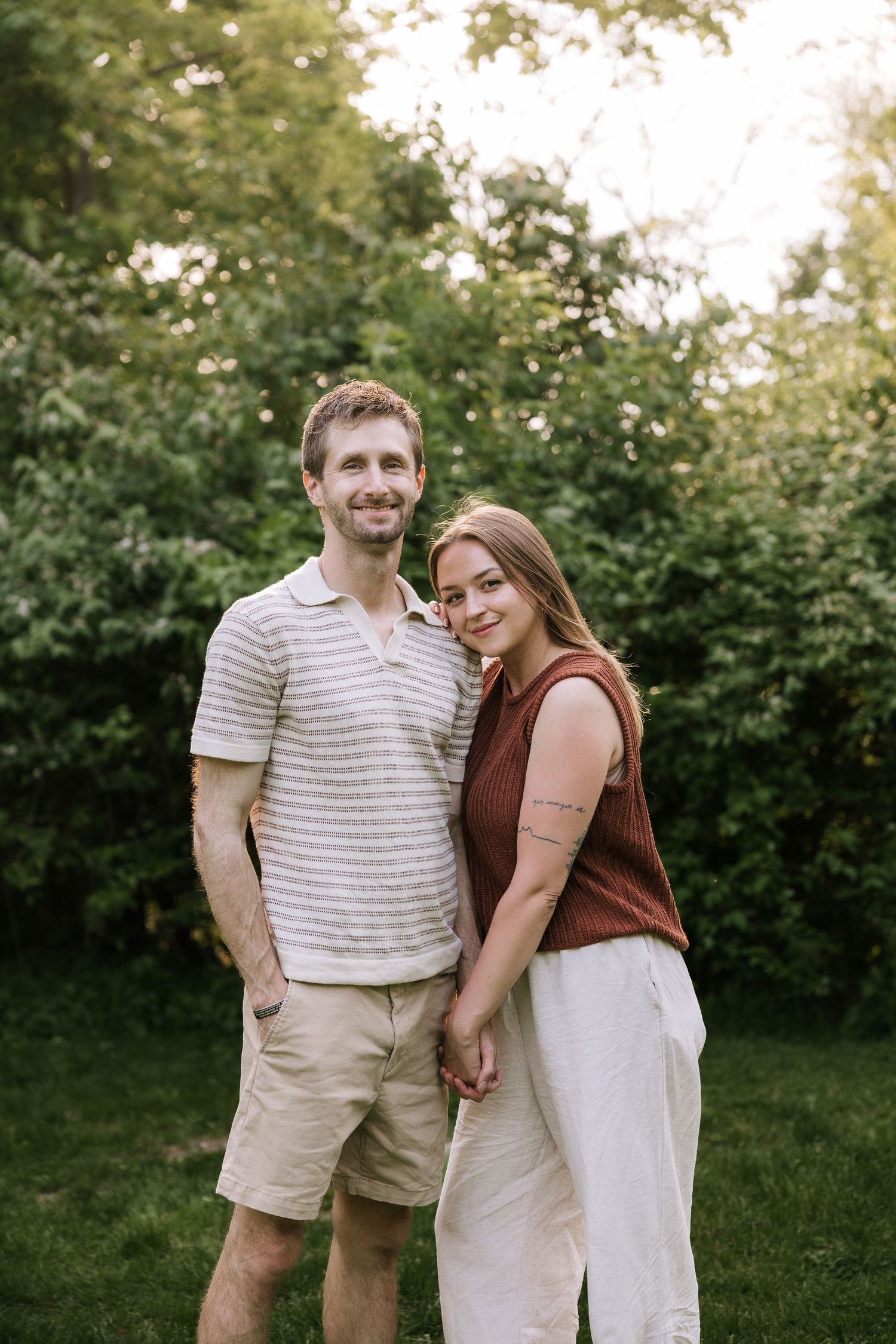 A smiling couple holds hands outdoors, man in striped polo and shorts, woman in rust top and white pants, green trees behind.
