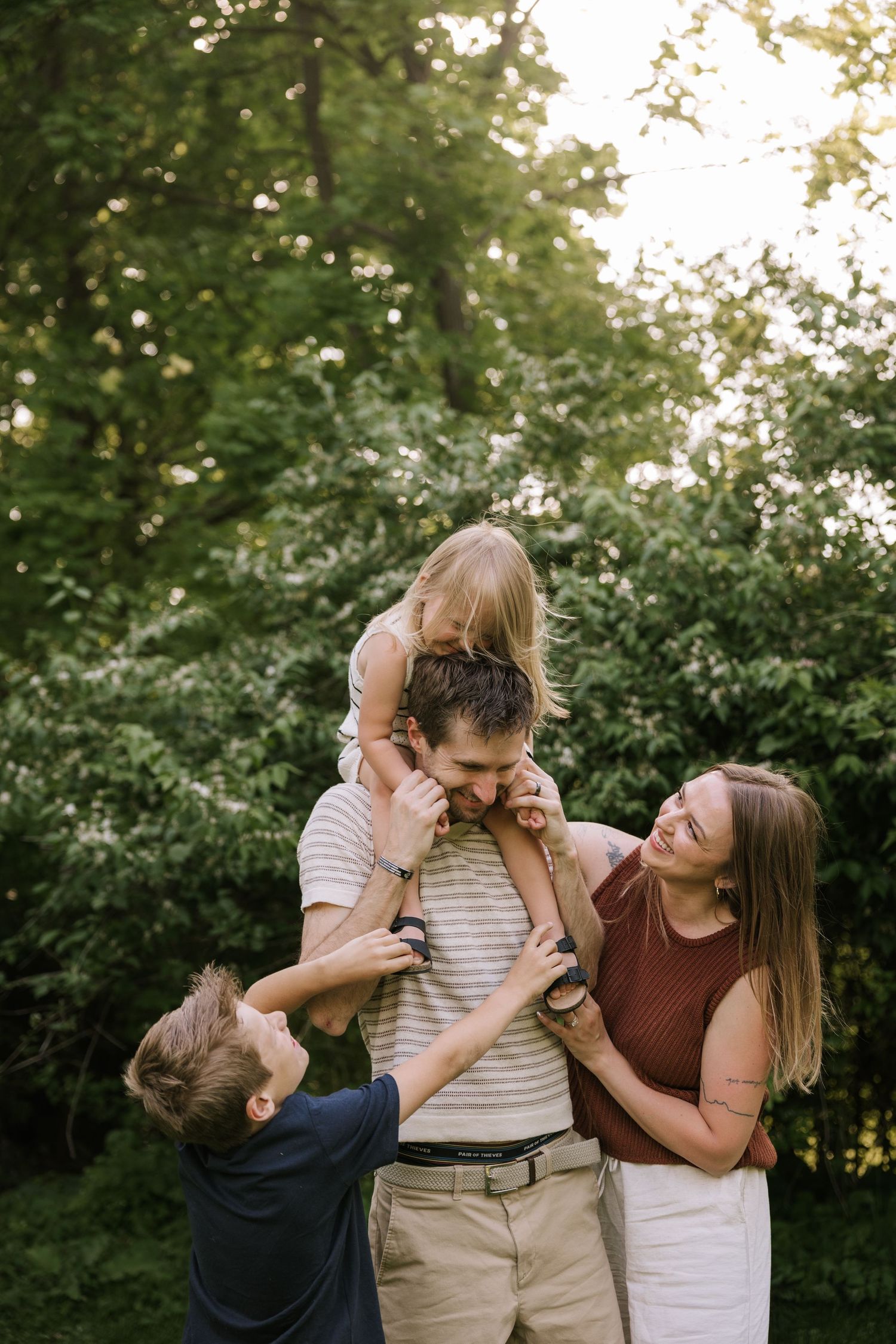 Happy family of four plays together outdoors, with dad carrying daughter on shoulders.