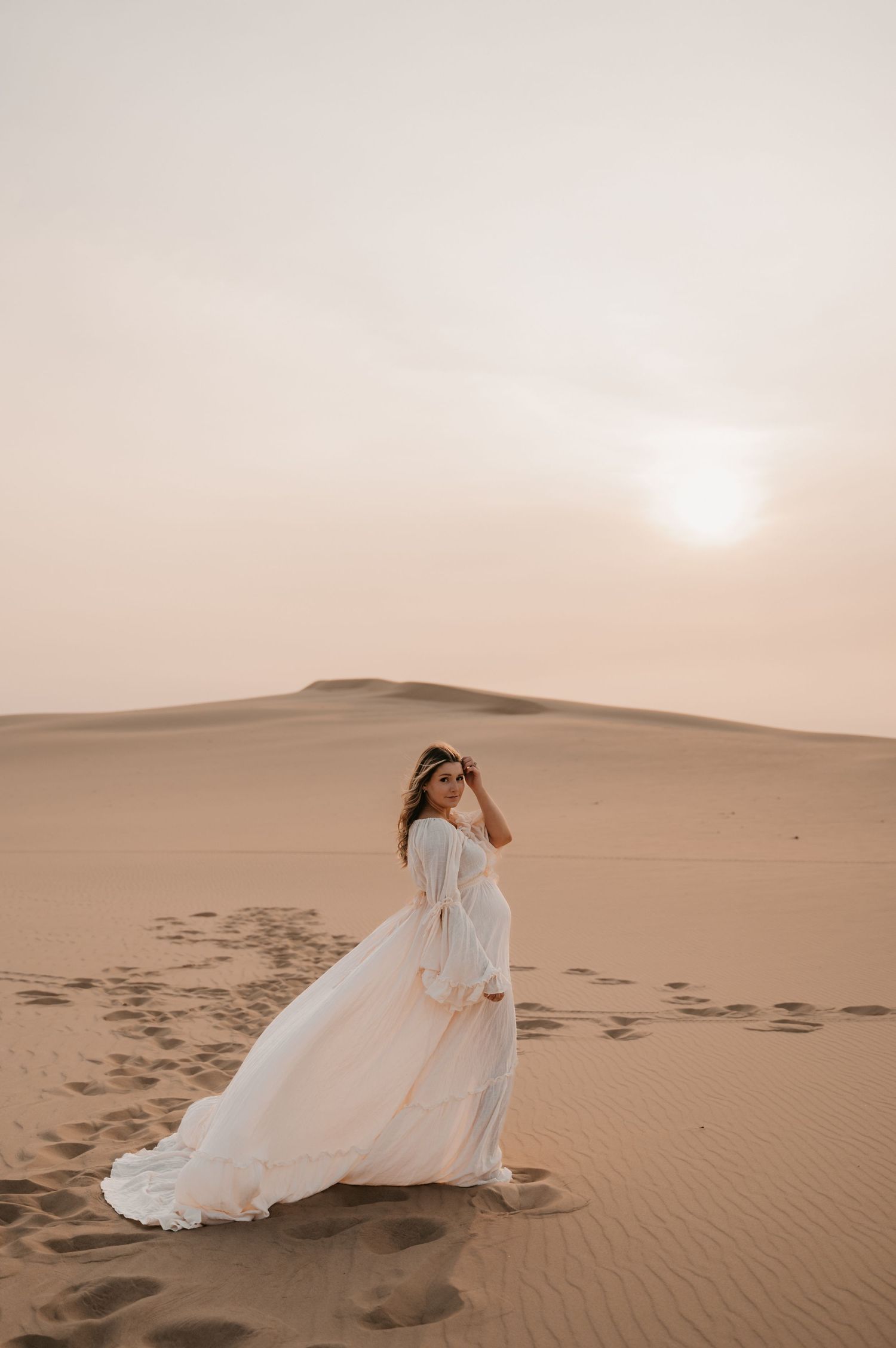 Woman in flowing white dress twirls on desert sand dunes at sunset.