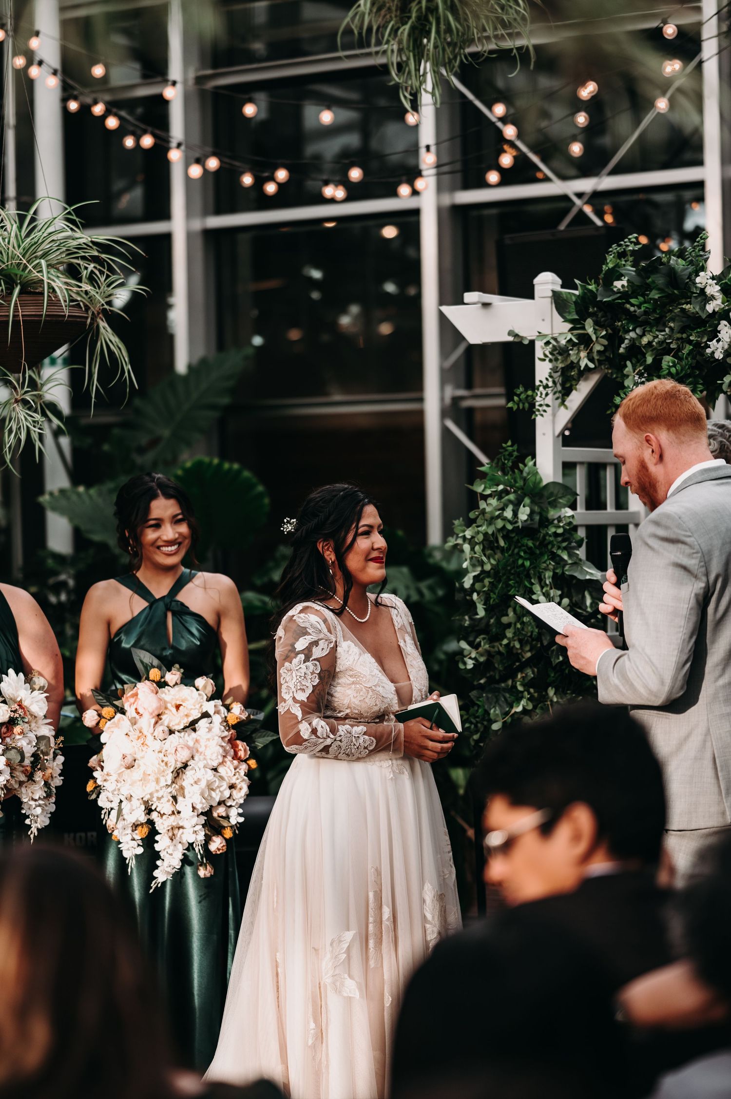 Wedding ceremony with bridesmaids in green dresses holding blush bouquets in greenhouse setting.