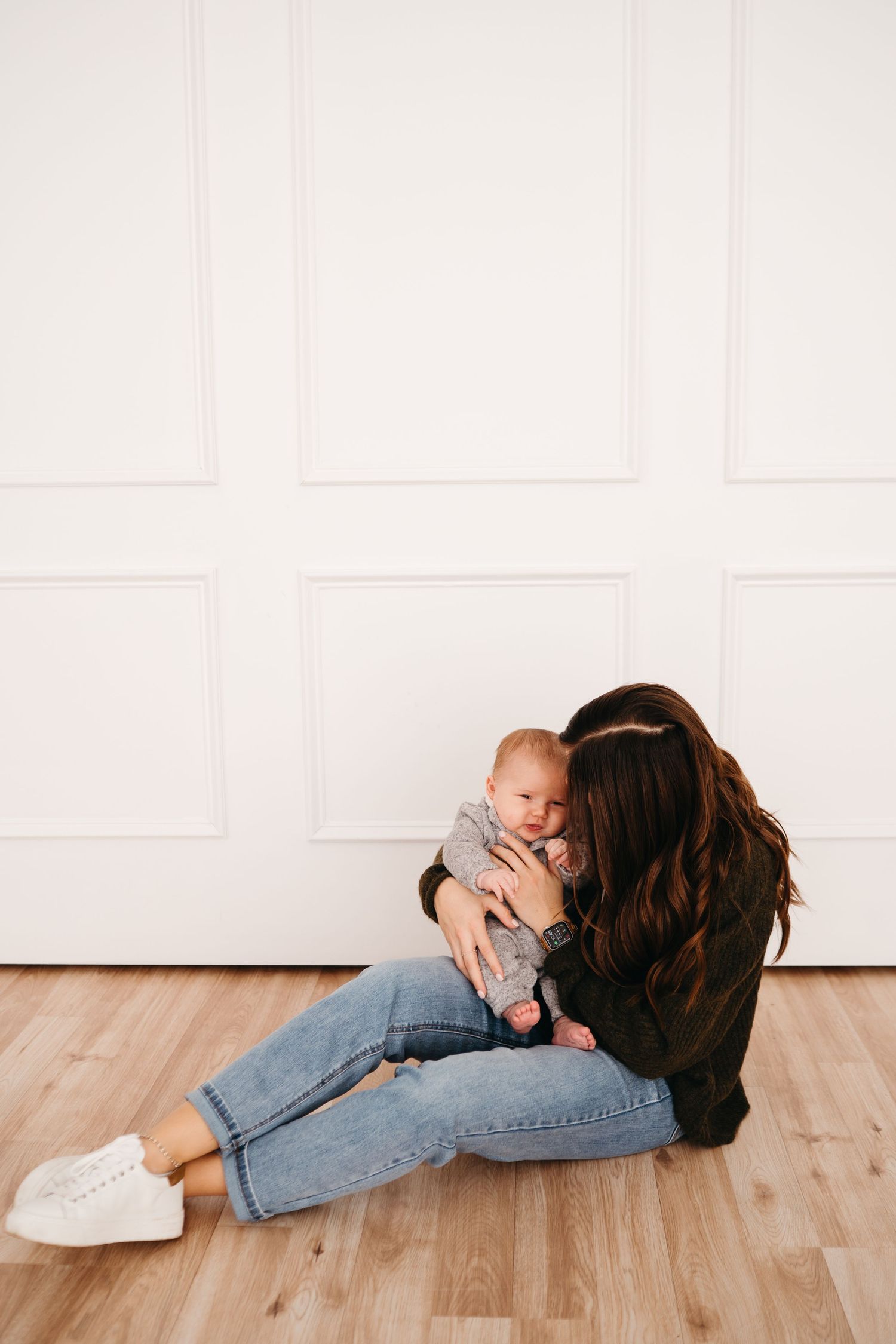 Parent sitting on wooden floor cuddling with baby against white wall.