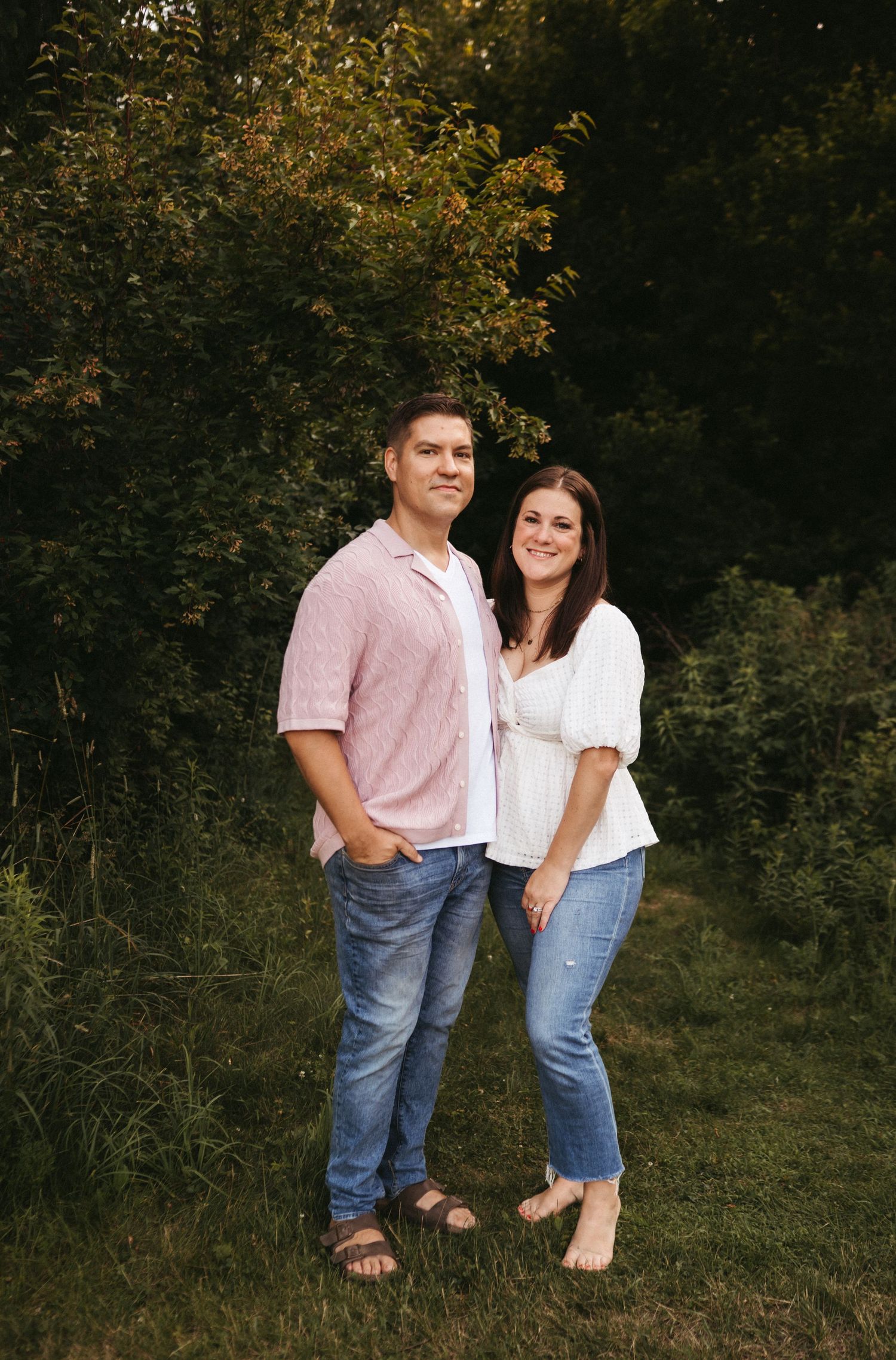 A couple stands together in casual attire against a wooded outdoor background.