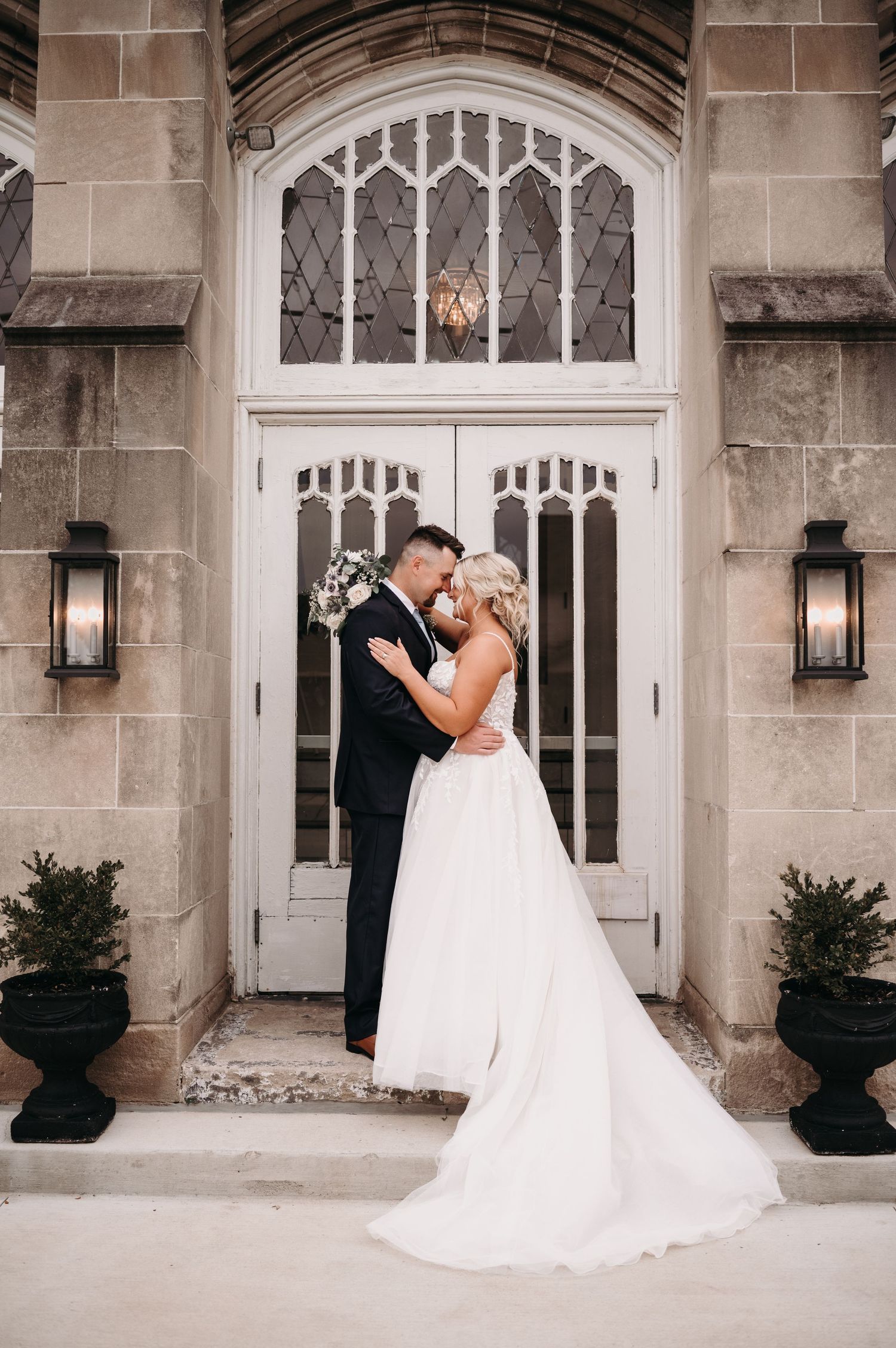 Romantic wedding couple embraces in front of elegant Gothic doorway with lanterns and white architecture.
