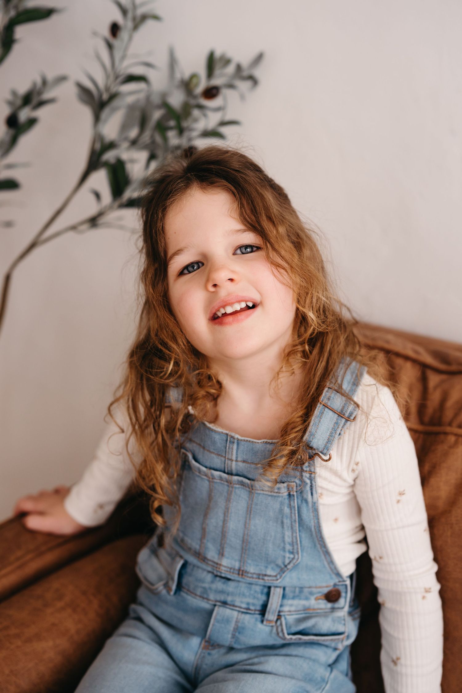 Young child wearing denim overalls and white long sleeve shirt sits on brown sofa with decorative foliage in background.