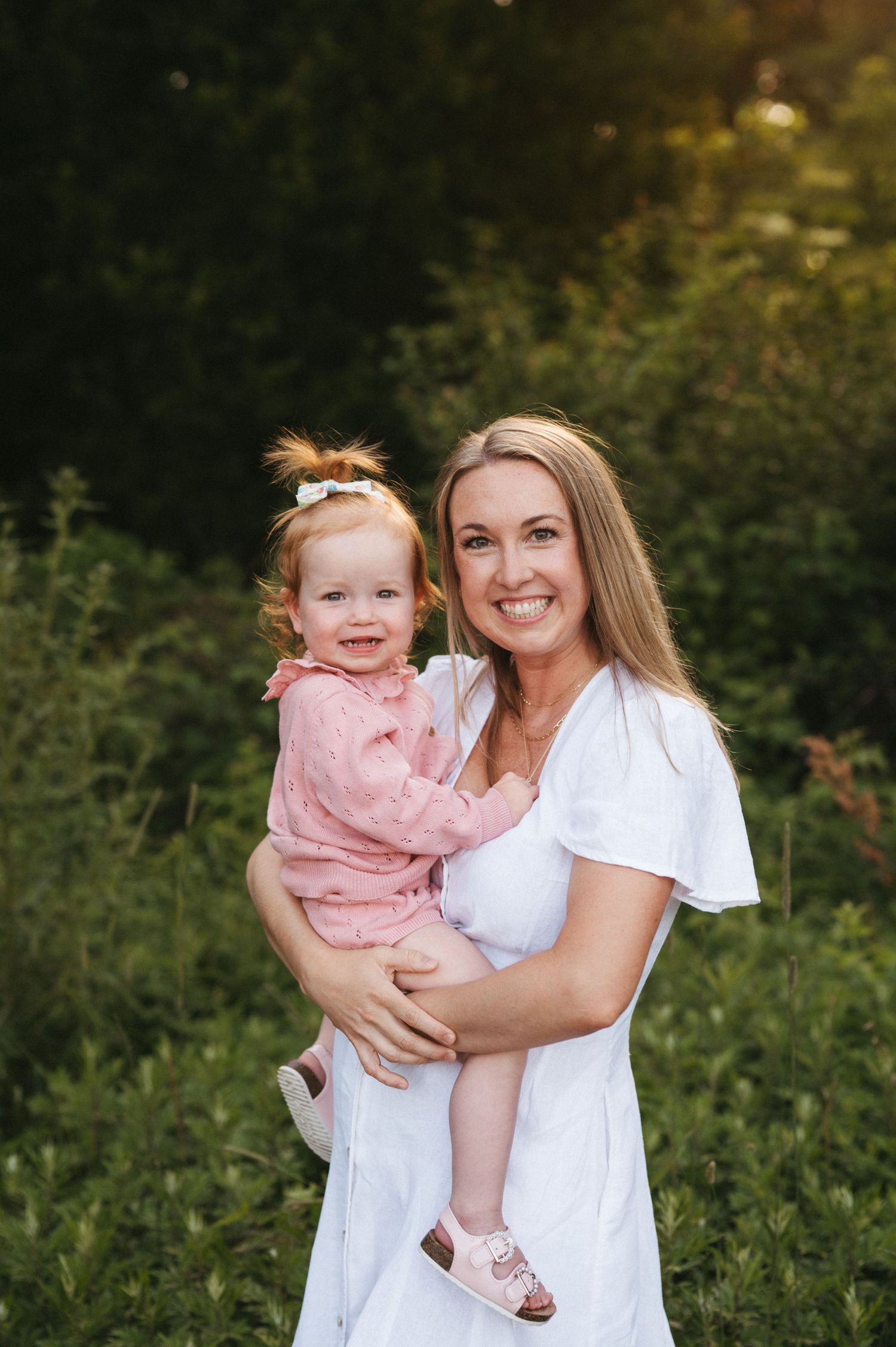 A person in white holds a small child wearing pink during an outdoor portrait session with natural greenery.