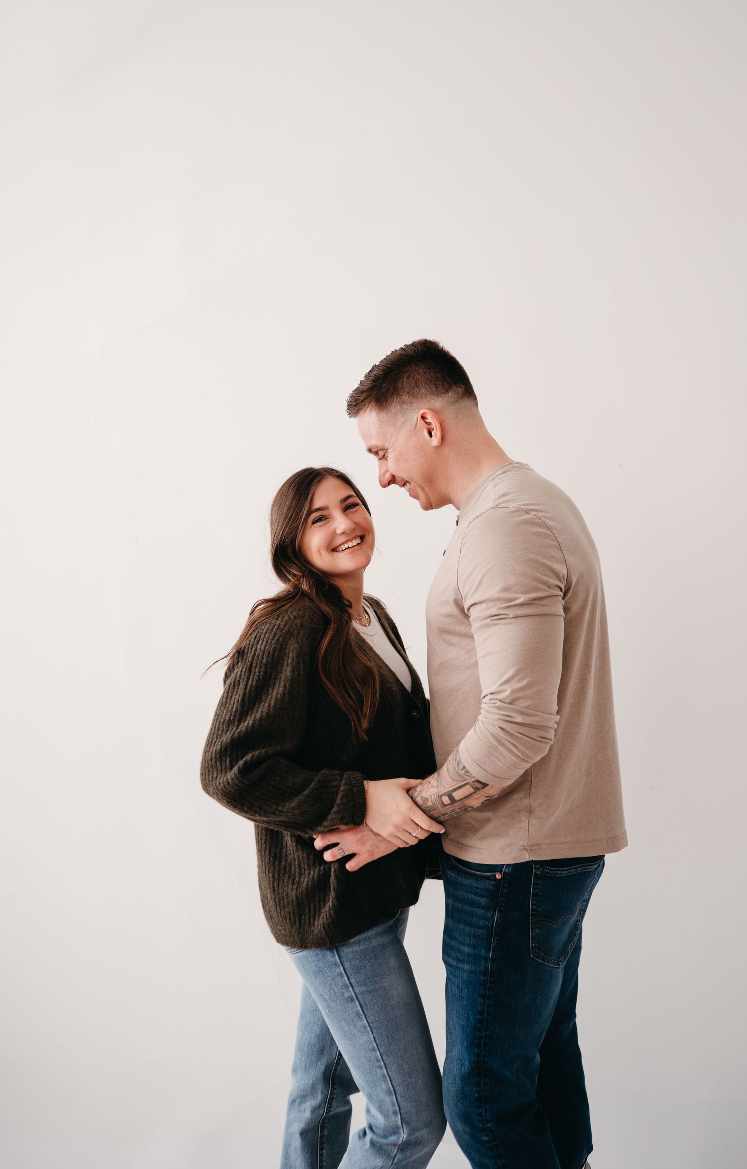 A couple shares a loving moment together against a white backdrop, wearing casual attire.