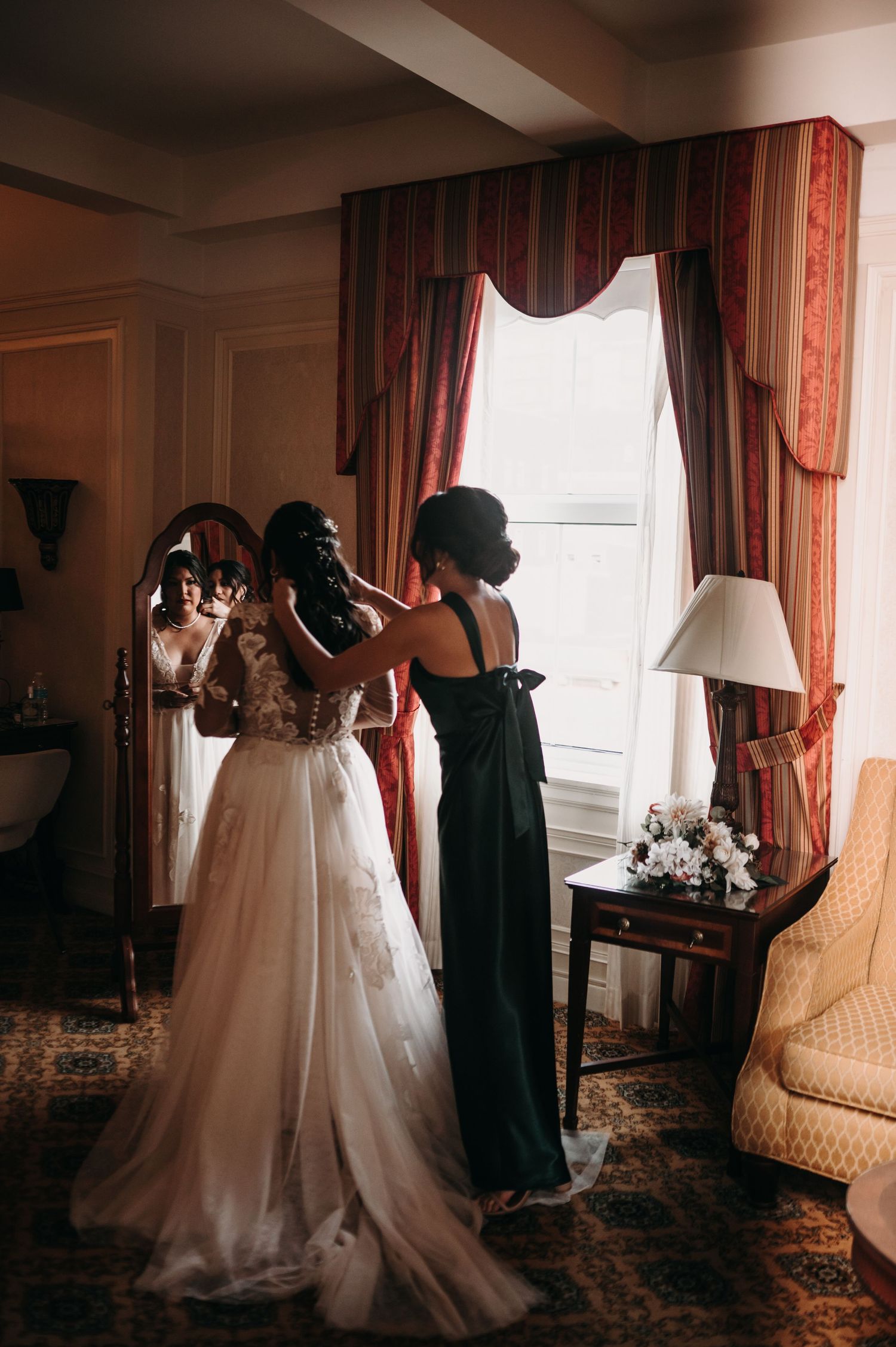 Final adjustments being made to a lace wedding gown in an ornate room with vintage furnishings.