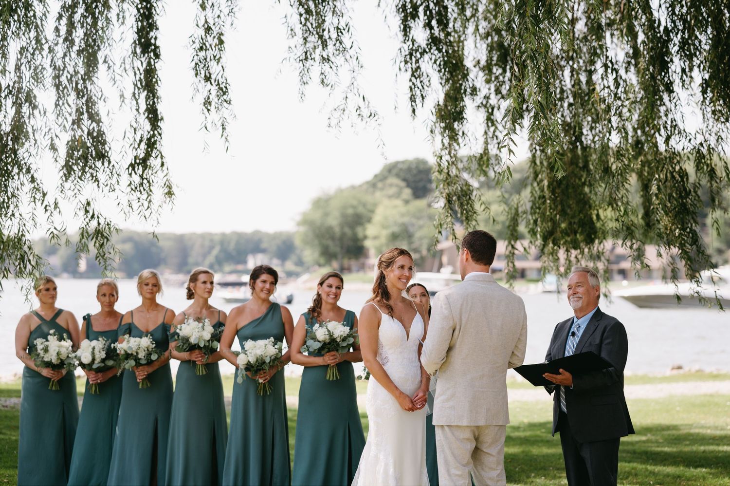 A wedding party stands beneath a weeping willow tree by a waterfront on a sunny day.