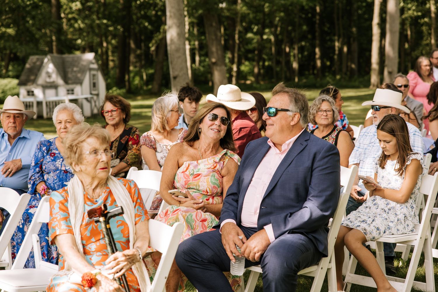 Wedding guests seated outdoors on white chairs during a garden ceremony beneath tall trees.