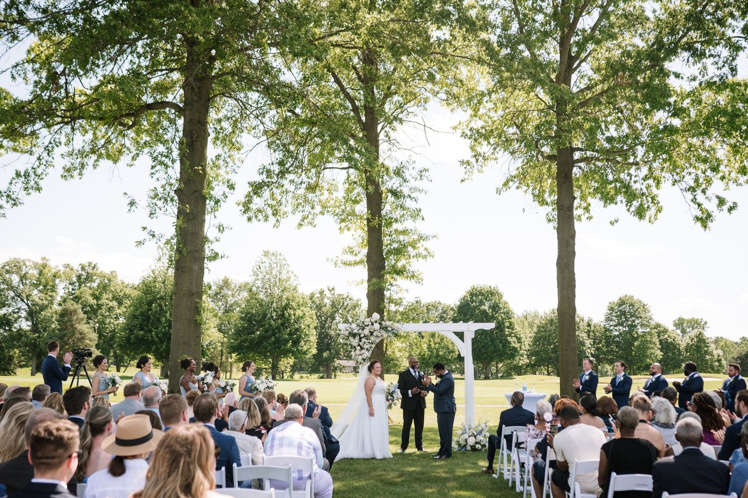 Outdoor wedding ceremony setup with white chairs arranged on a lawn under tall shade trees.
