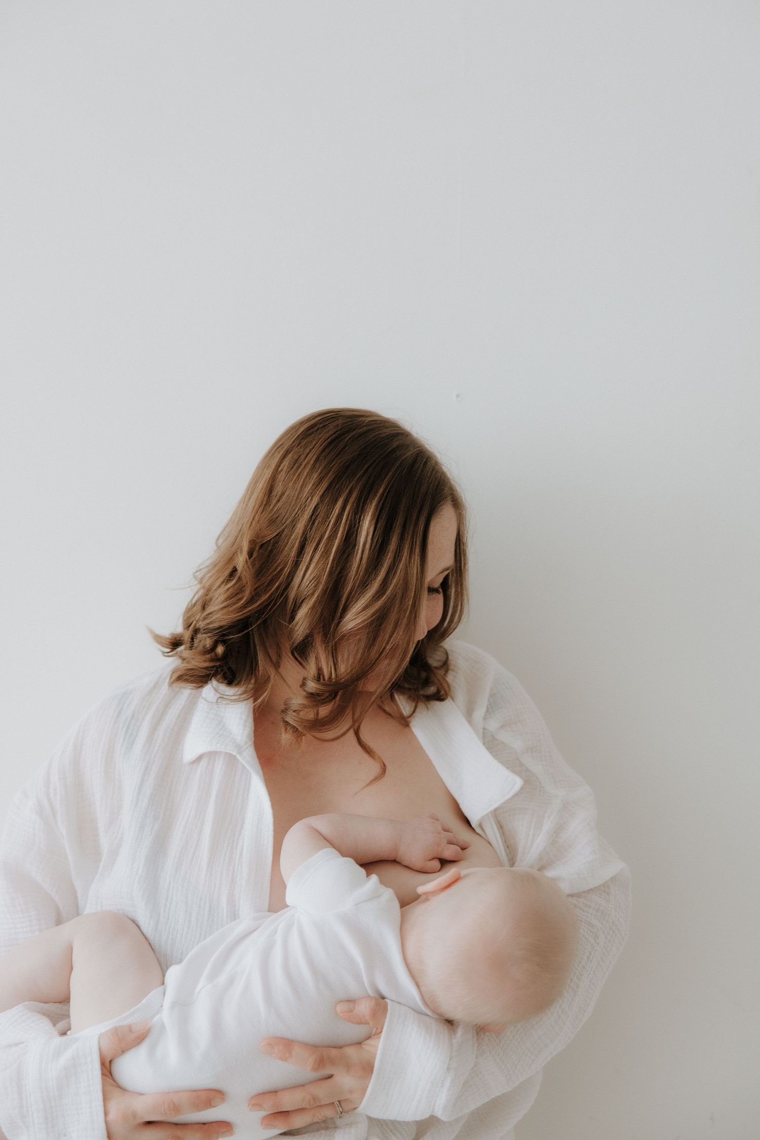 Mother with wavy hair in white linen shirt breastfeeding baby against white wall.