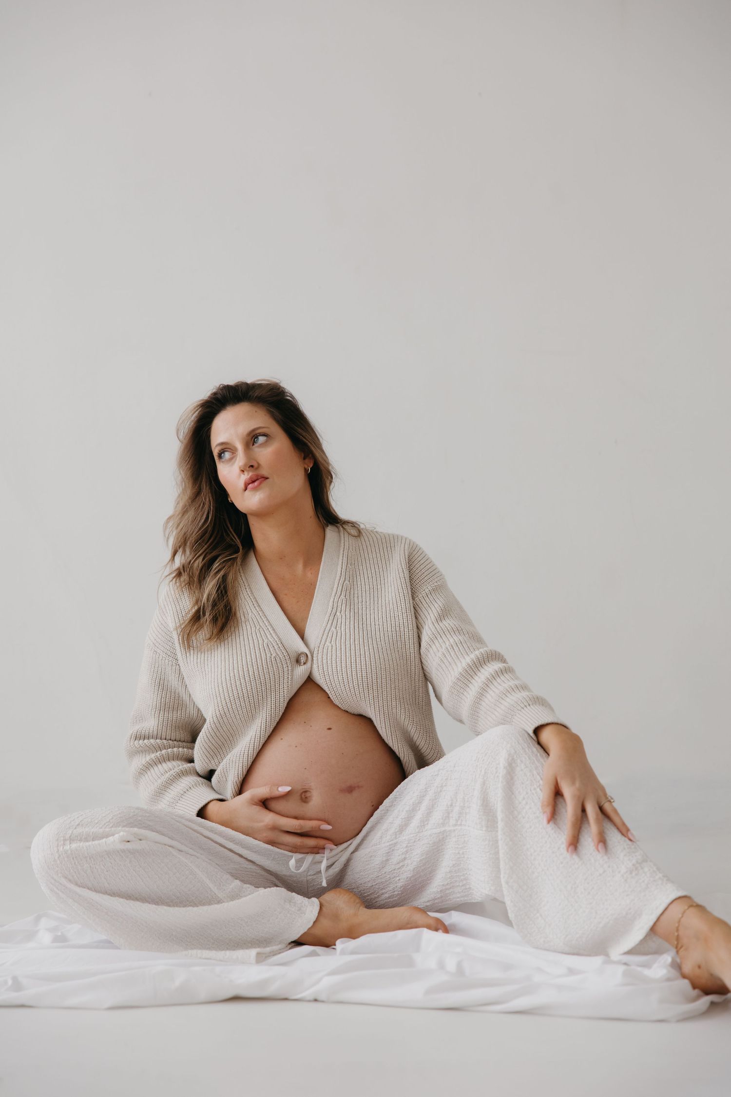Pregnant woman sits cross-legged on white floor cradling baby bump in neutral knit cardigan.