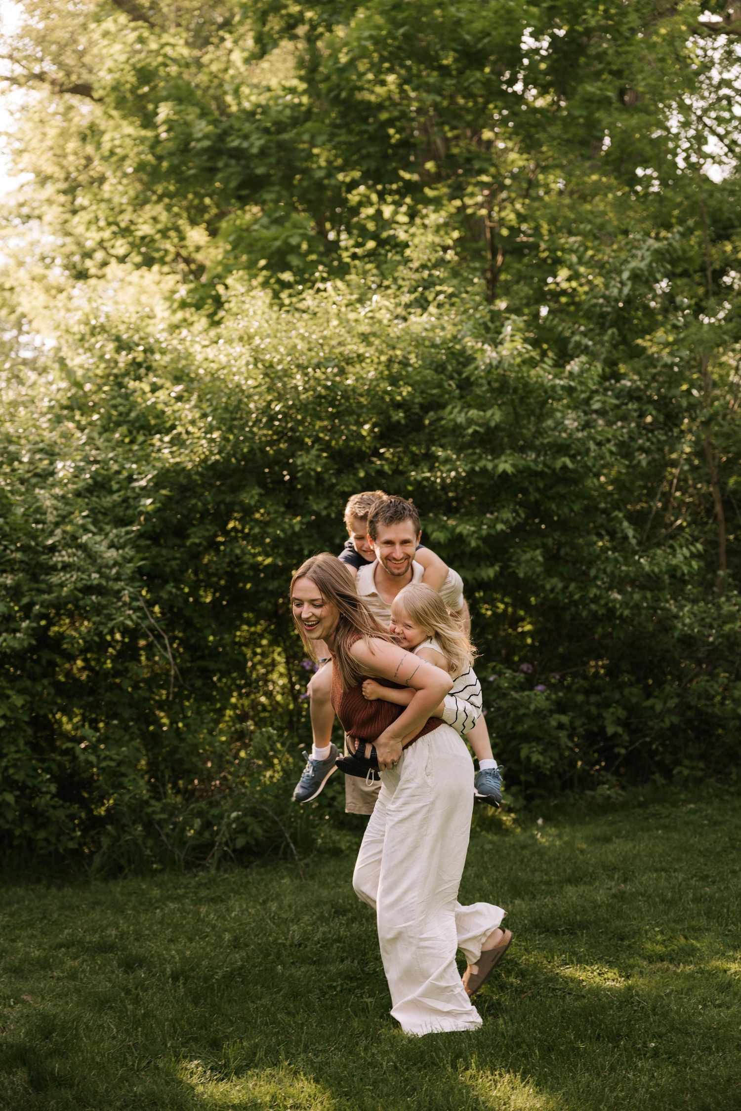 Happy family playing outdoors, parents giving children piggyback rides on lush green lawn surrounded by trees.