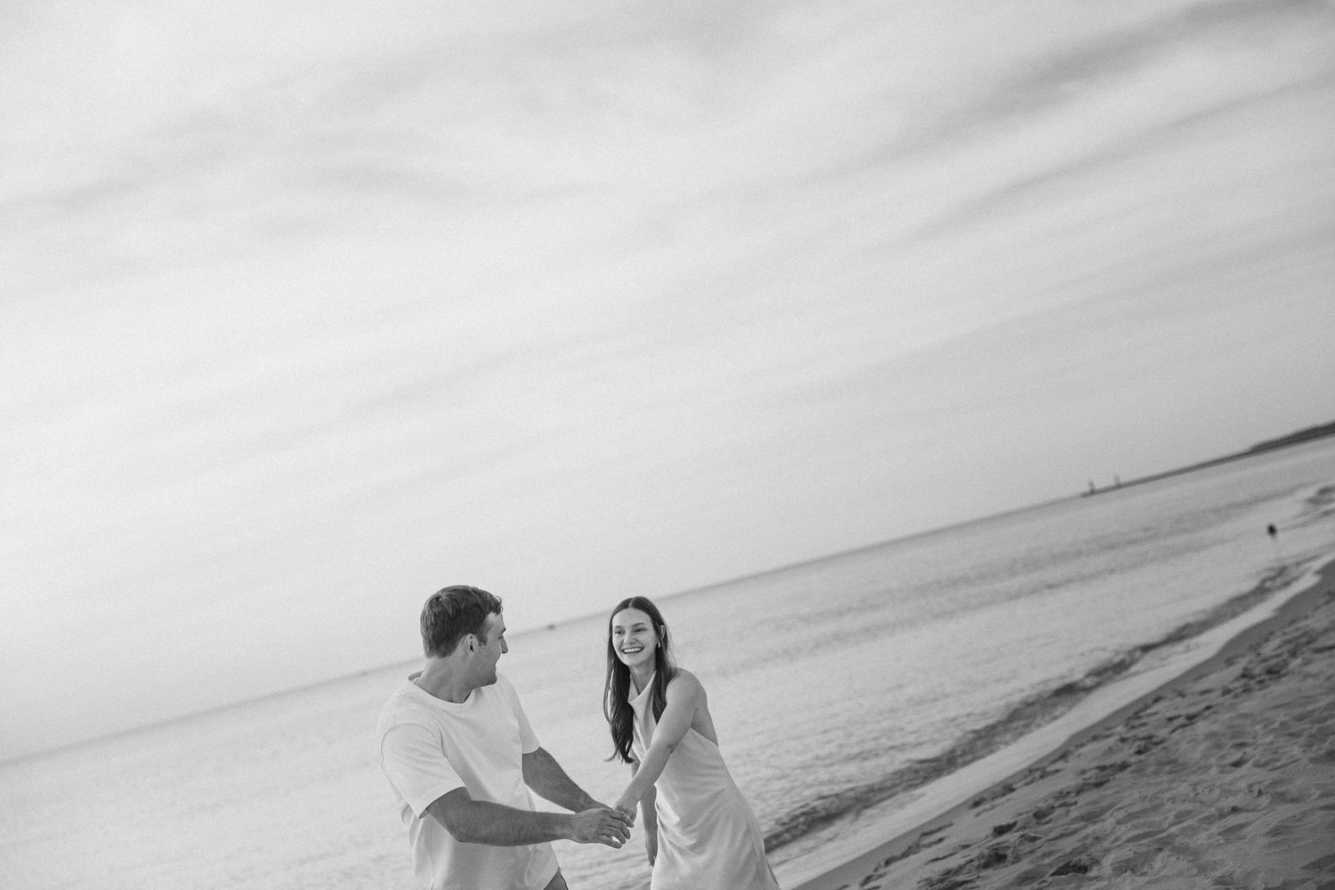 Black and white photo of a couple holding hands and laughing together on a sandy beach by the ocean.