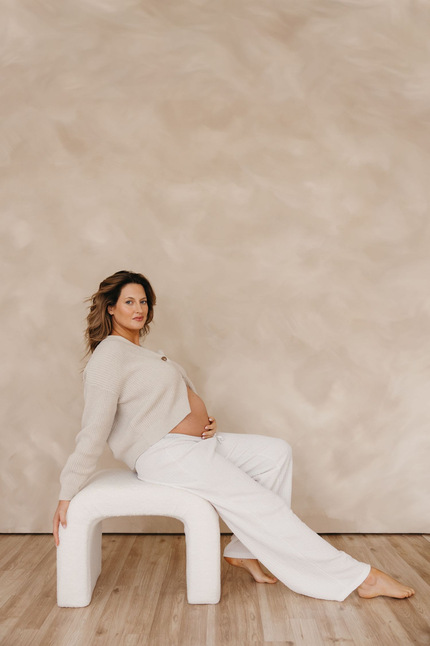 Pregnant woman in beige top and white pants sitting on white stool during elegant maternity shoot.