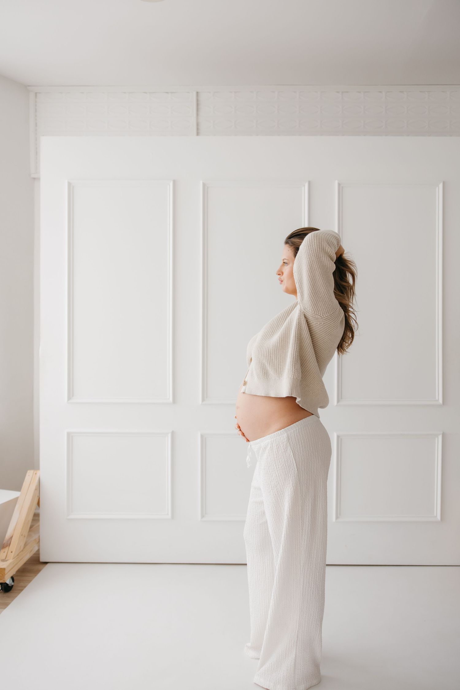 Pregnant woman in white crop top and pants standing sideways, showing baby bump against white paneled wall.