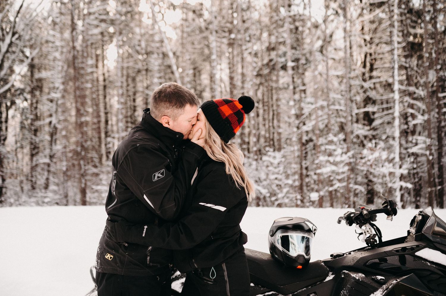 Two people in black winter clothing embrace near a snowmobile surrounded by snow-covered trees.