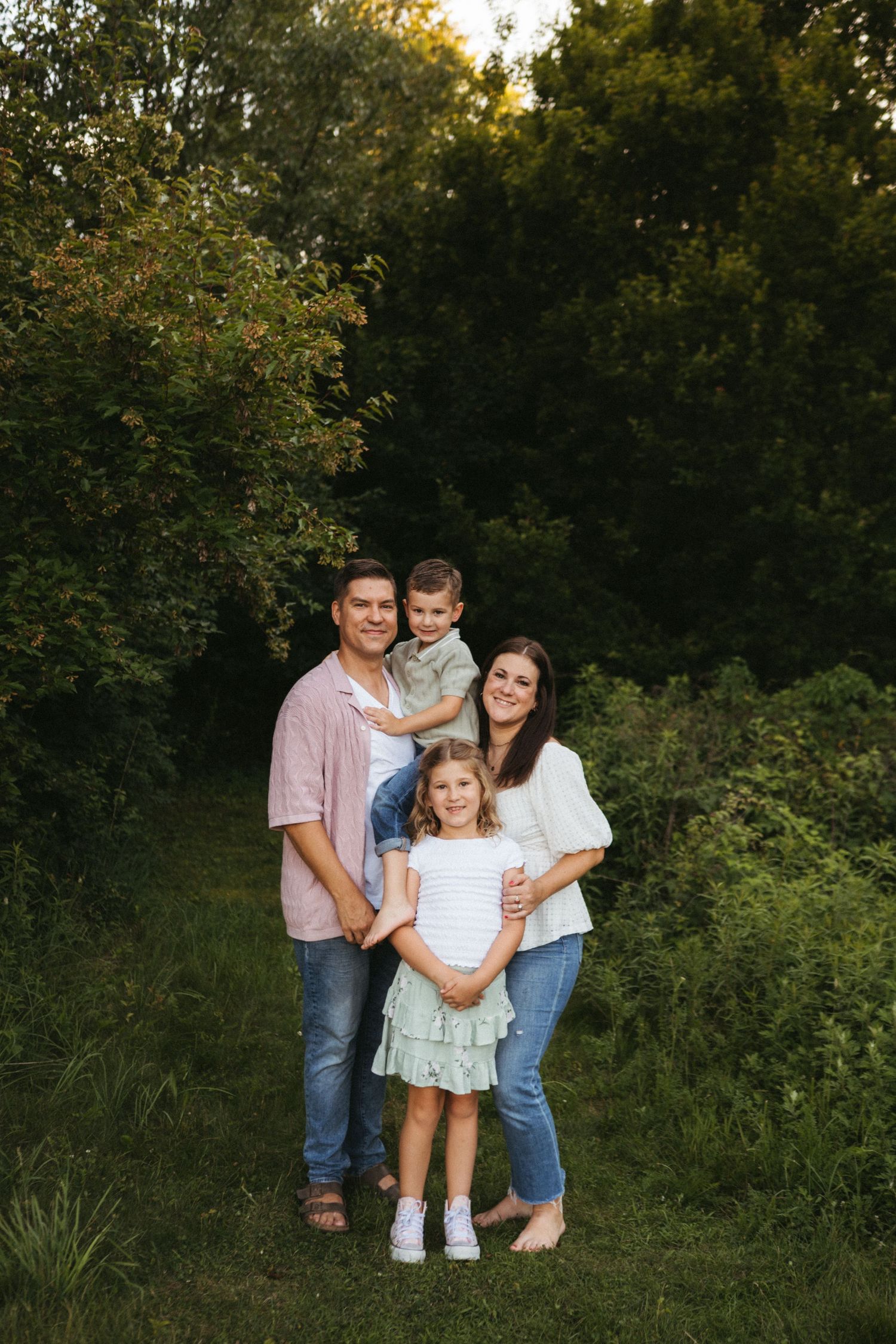 A family of four poses together for a portrait in a lush green outdoor setting at sunset.