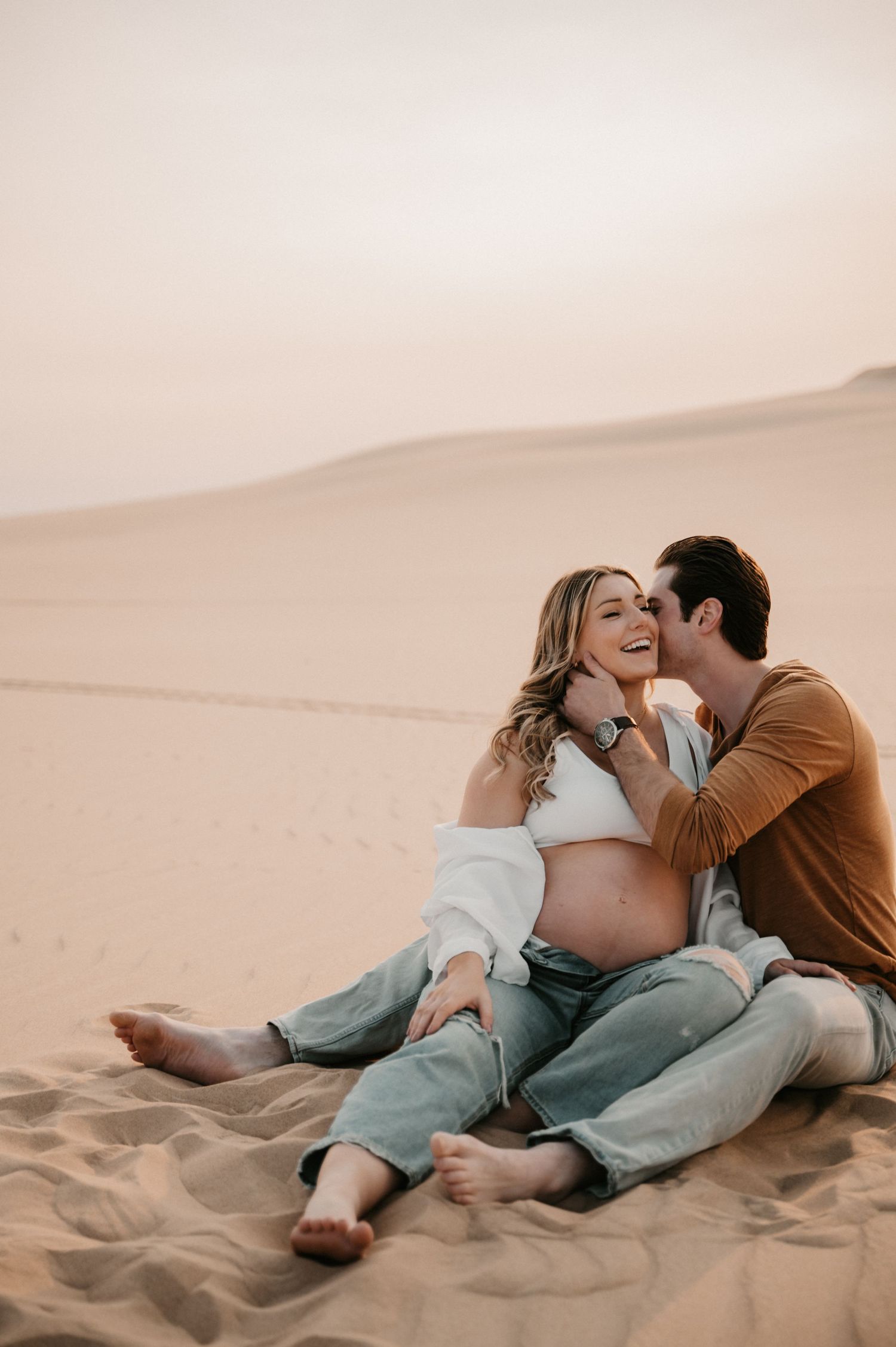Candid maternity moment captured between couple sitting on desert sand during golden hour.
