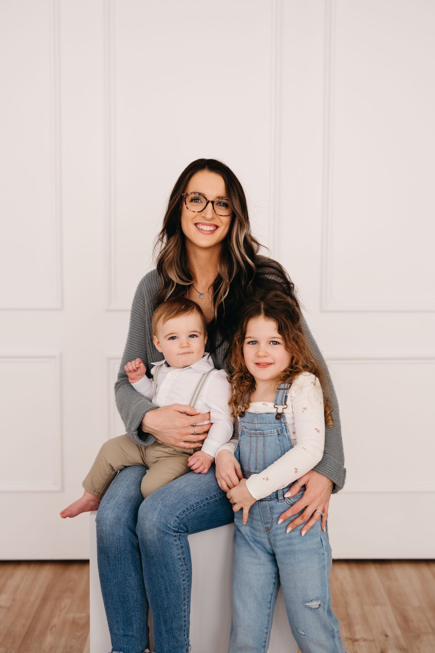 A person wearing glasses sits with two young children in casual denim outfits against a white wall background.