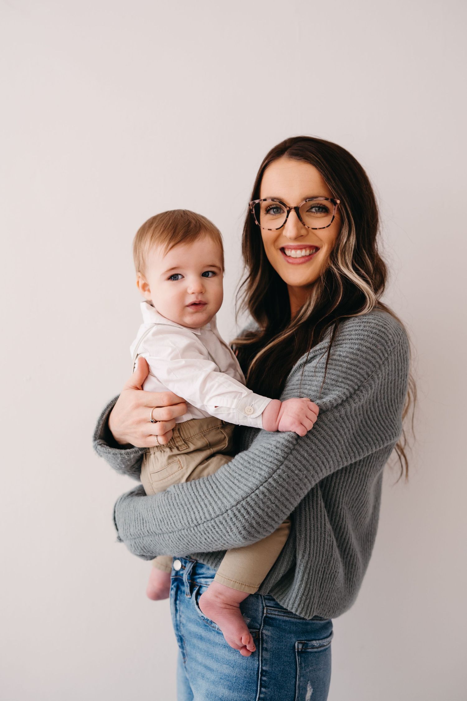 A parent wearing glasses and a grey sweater holds their smiling baby in a white shirt and khaki pants against a neutral backdrop.