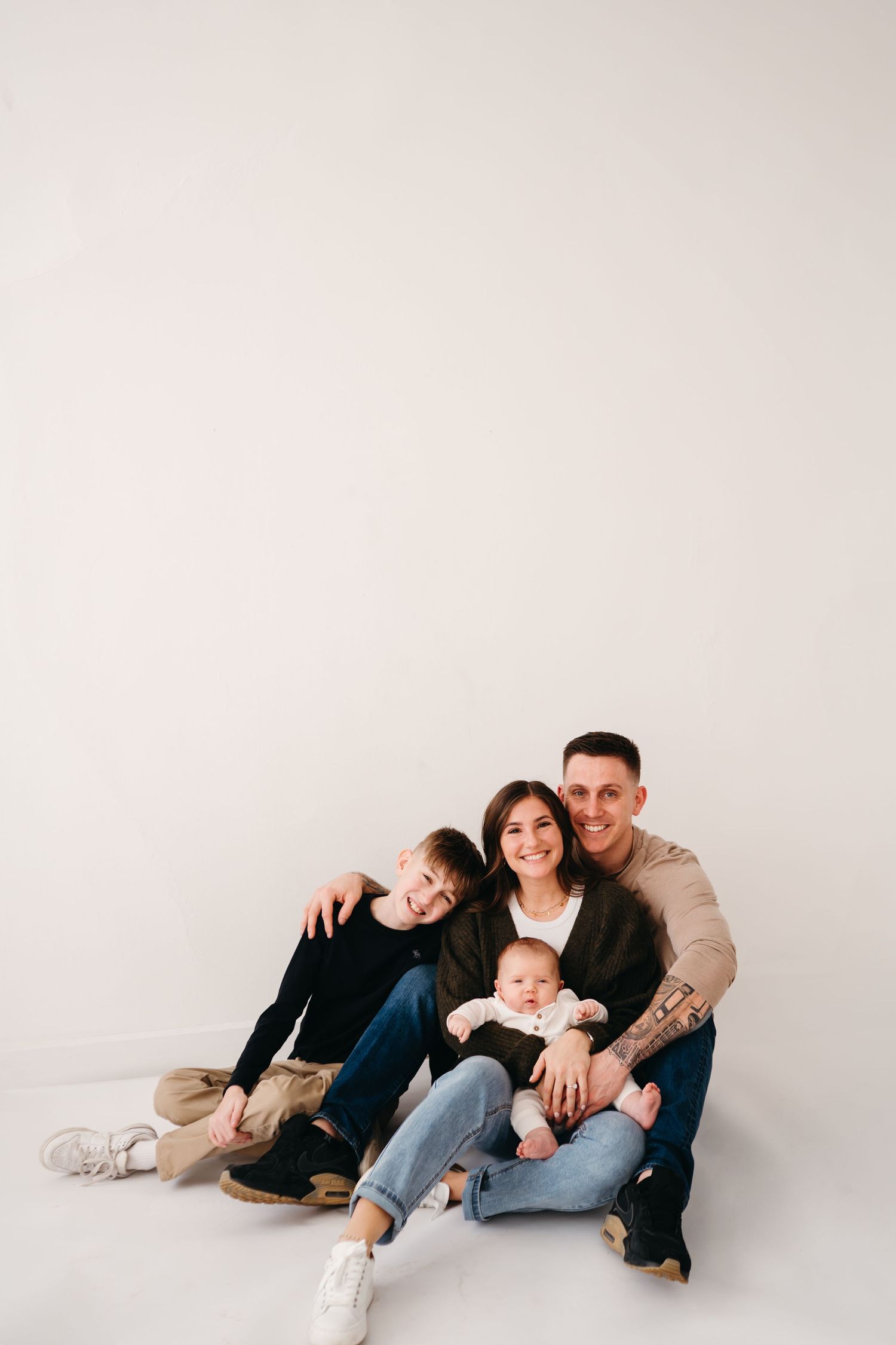 A casual family portrait of four sitting together on a white floor in coordinated outfits.