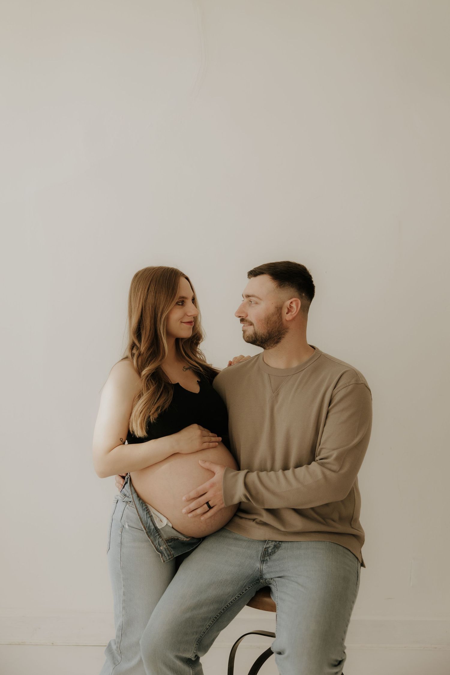 Tender maternity moment captured as couple shares intimate pose on studio stool.