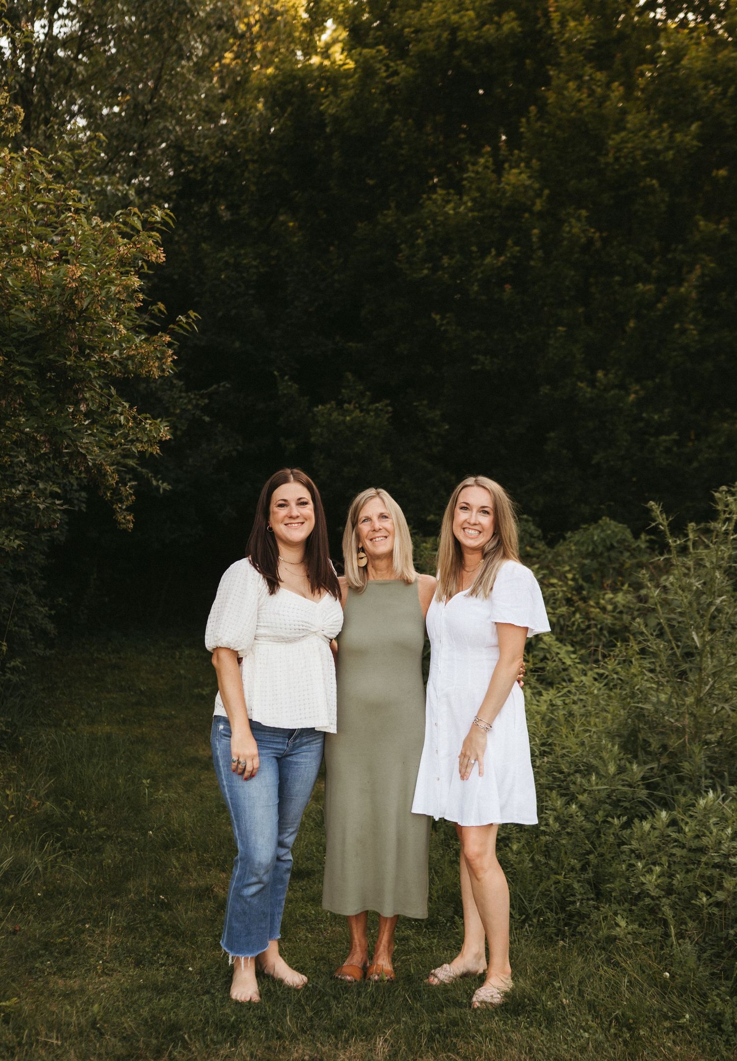 Three women stand together outdoors wearing casual summer outfits against a wooded backdrop.