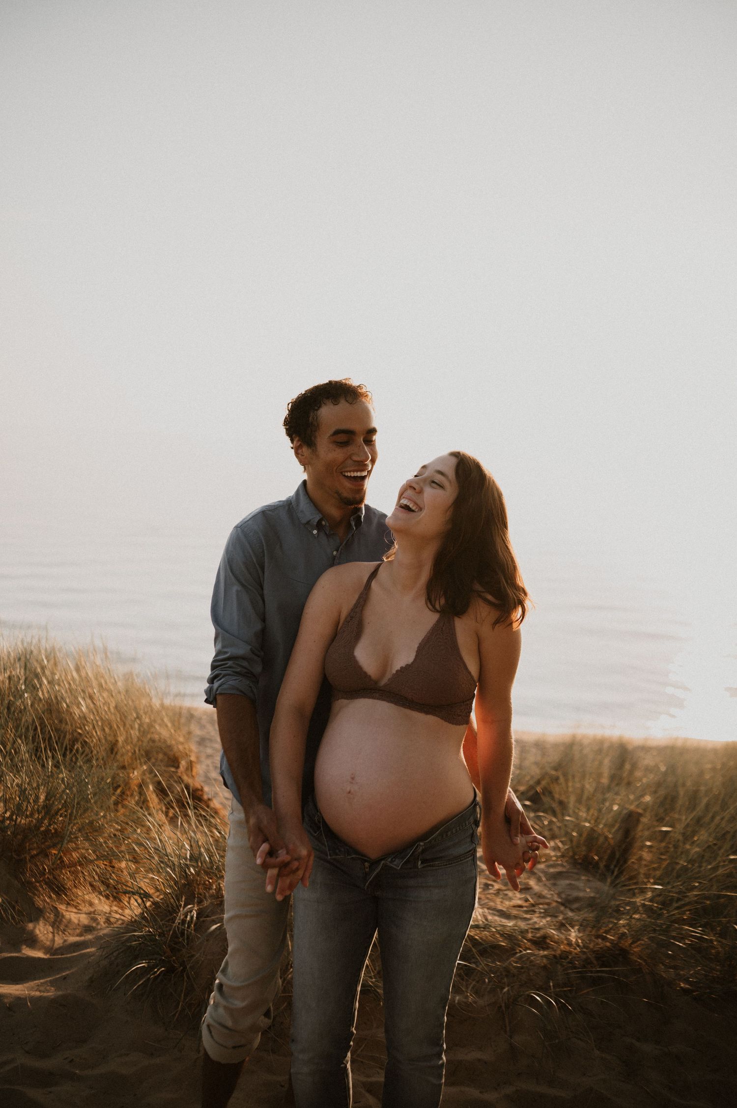 Joyful maternity photo of couple laughing together at sunset by the ocean.