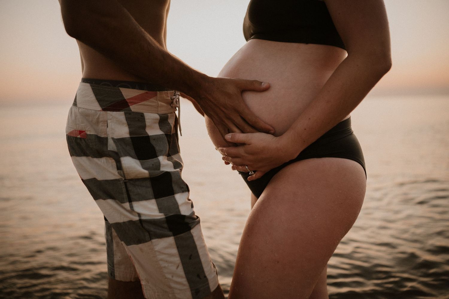 Intimate maternity photo of a couple embracing on a beach at sunset with waves in the background.