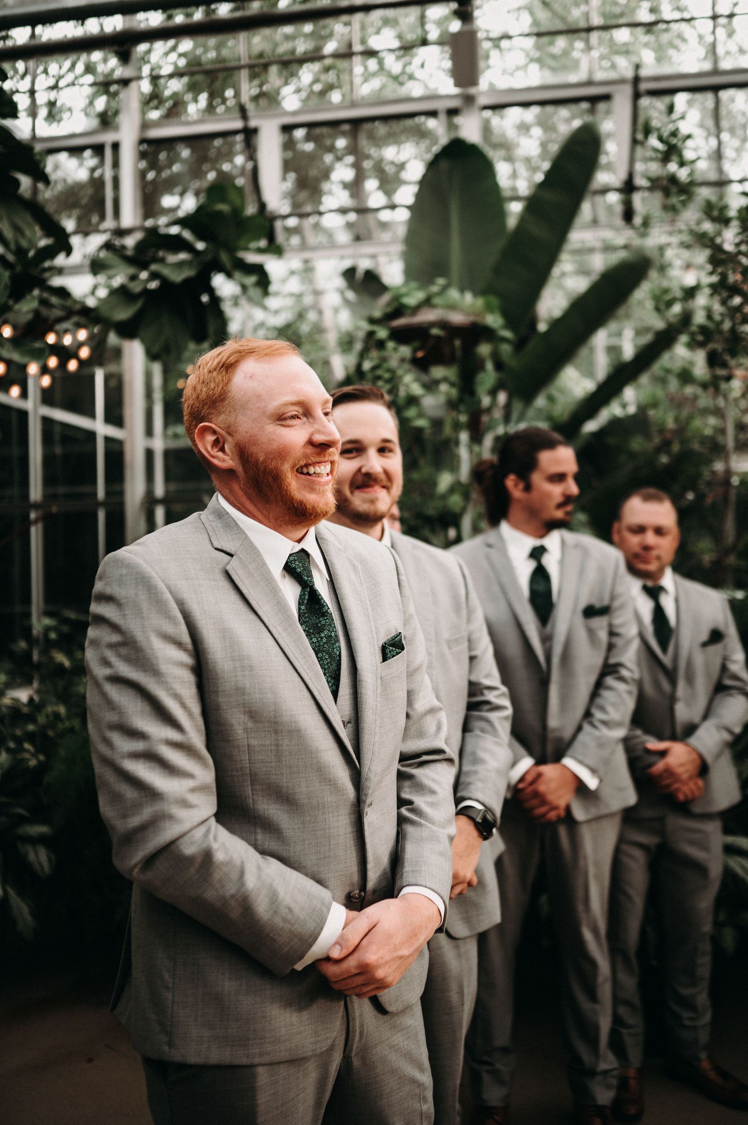Groomsmen in matching gray suits and ties stand together in a greenhouse setting with tropical plants.