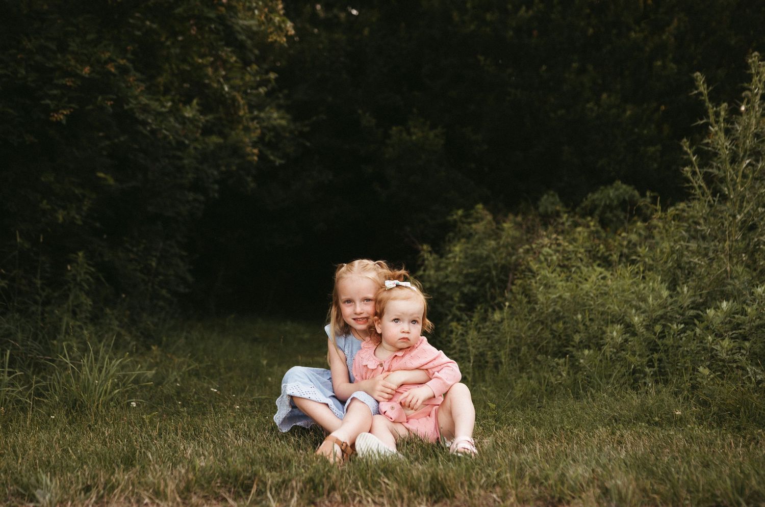 Two children sitting together on grassy hillside in summer with trees in background.