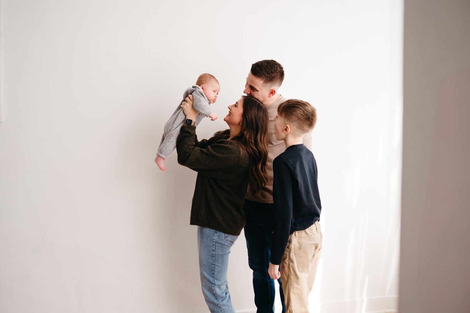 A small baby is lifted up high by family members against a white wall background.