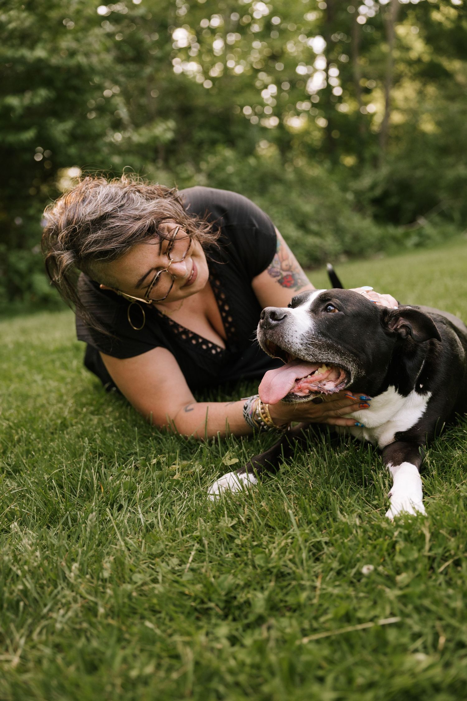 Tattooed woman with glasses cuddles a black and white dog while lying on green grass.