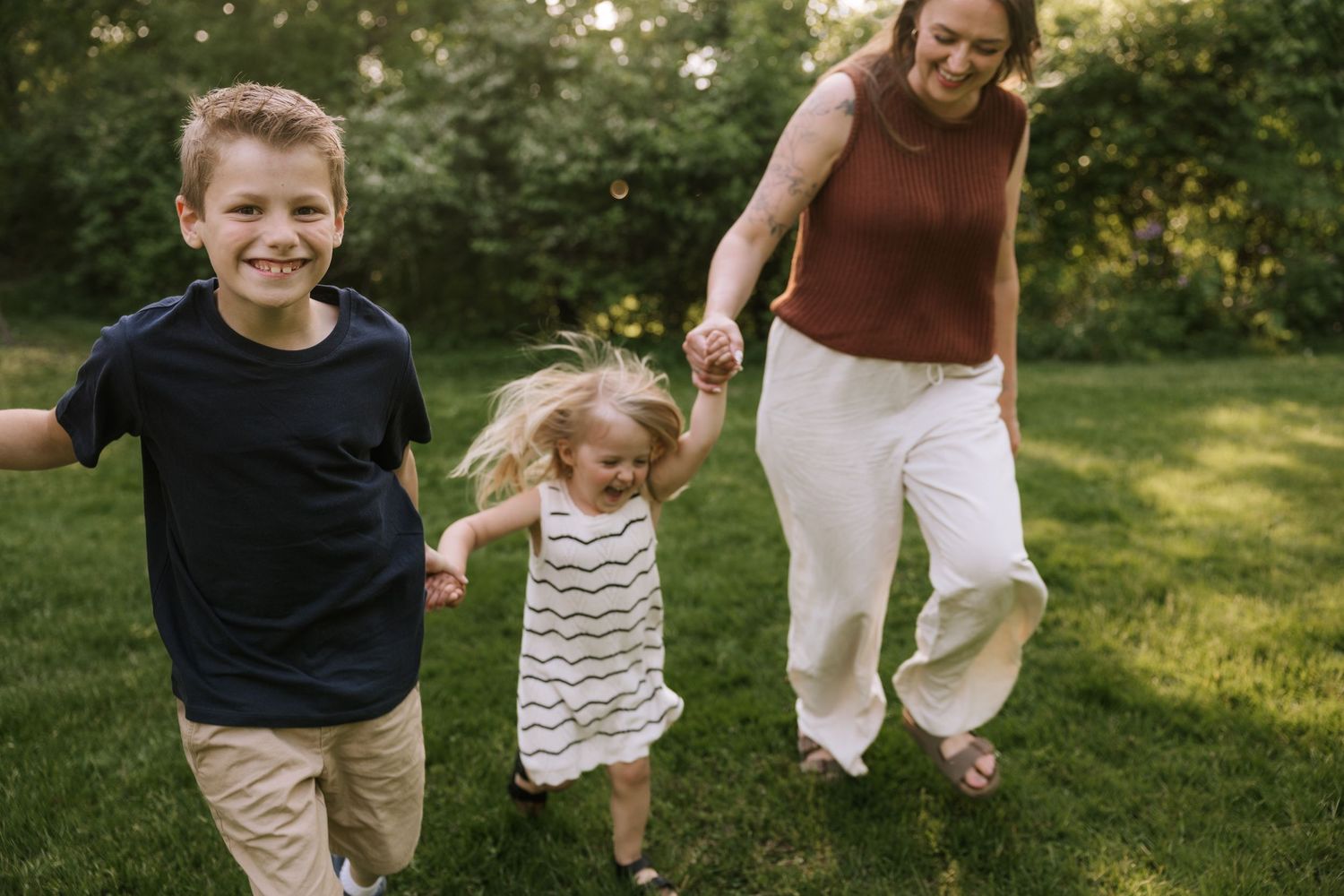 A joyful mother and two young children run and play together on a lush green lawn outdoors.