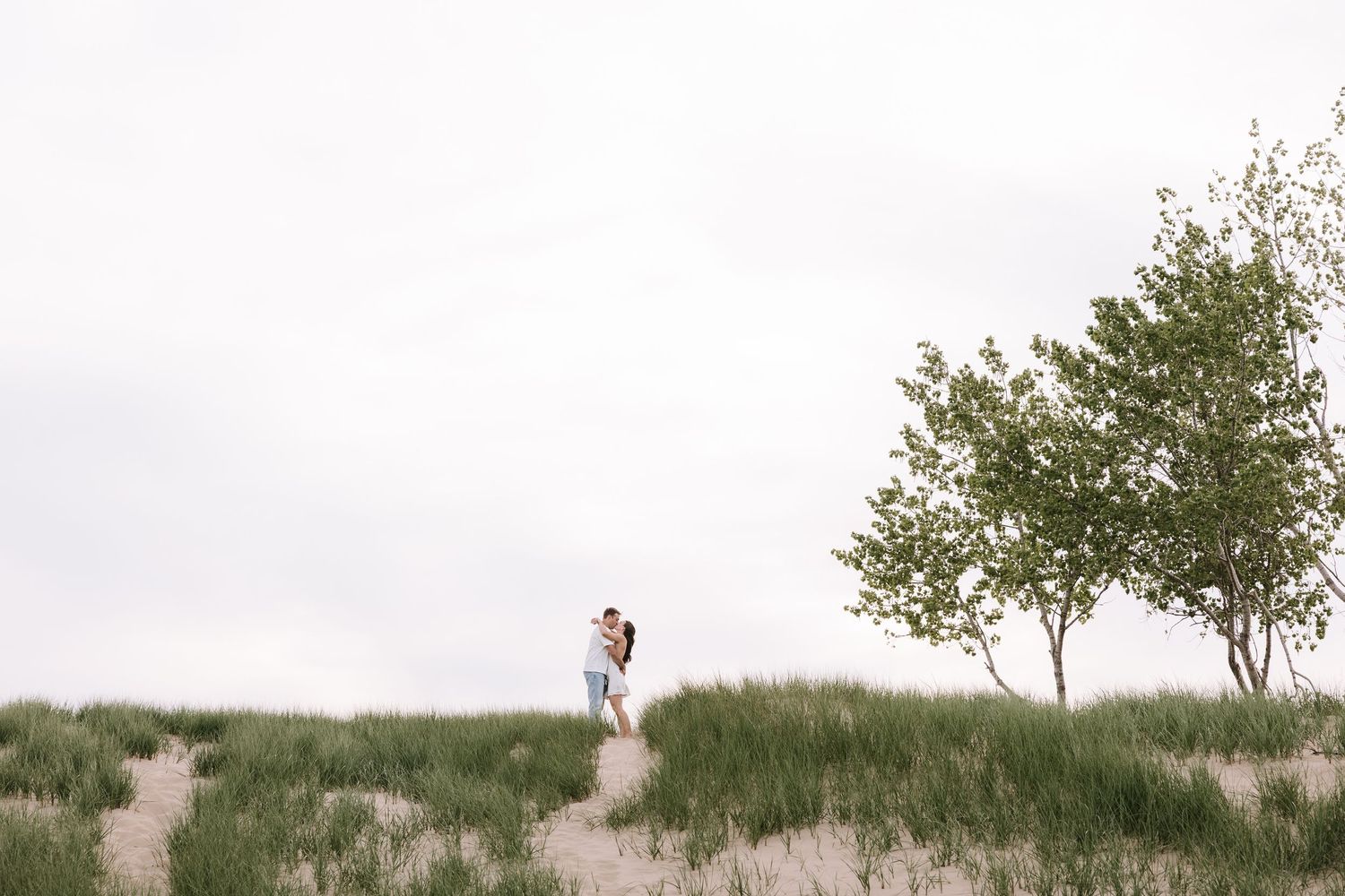 Couple embracing on sandy dunes with green grass and trees under overcast sky.