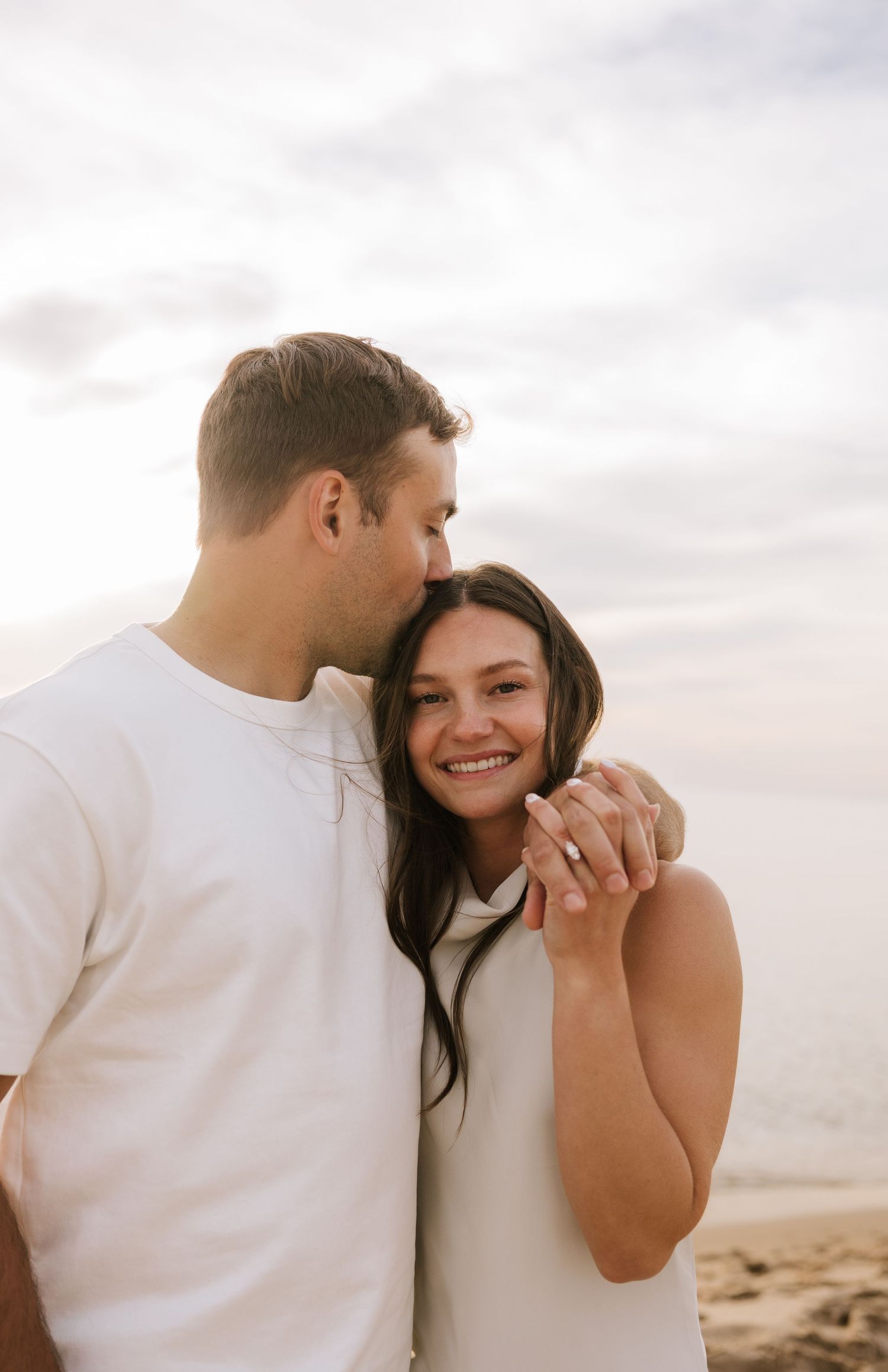 Man kissing woman on cheek at sunset beach, woman smiling and showing engagement ring.