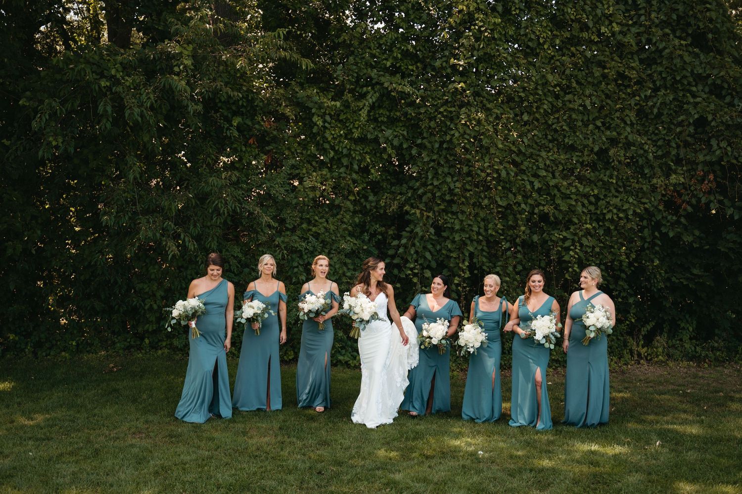 Wedding party in light blue dresses and dark suits poses together on grassy lawn at outdoor celebration.