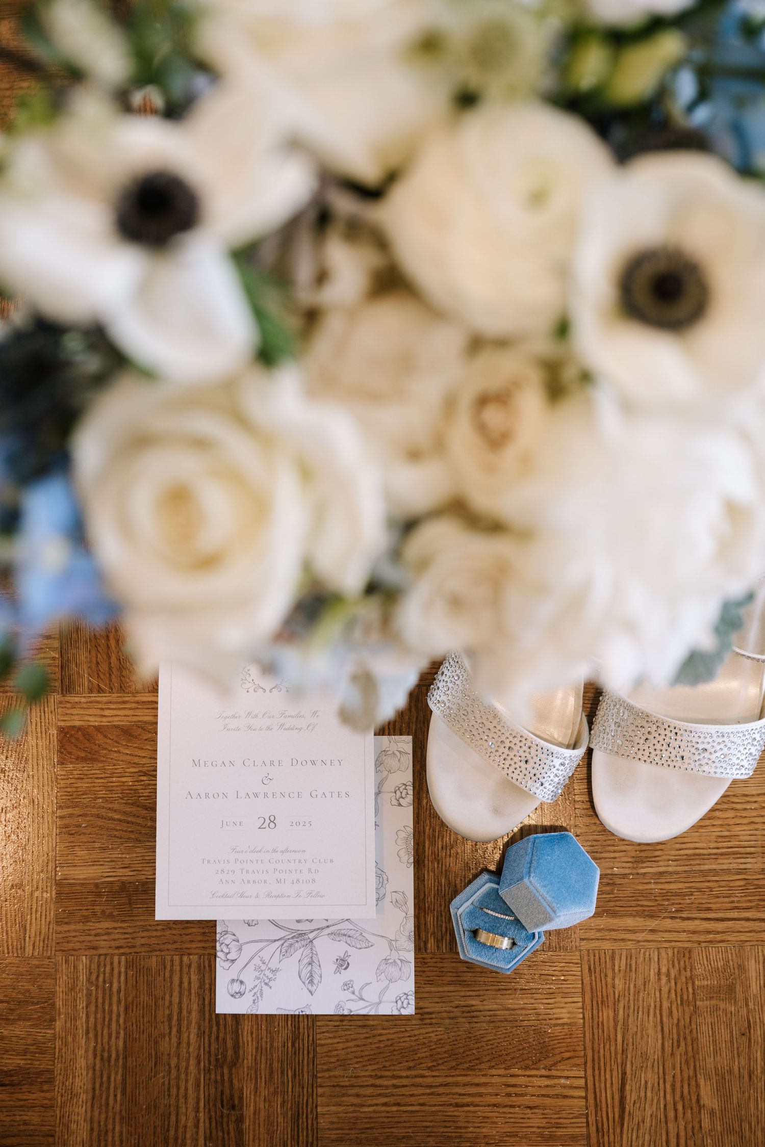 Wedding details including white roses and blue ring box arranged on wooden surface with invitation cards.
