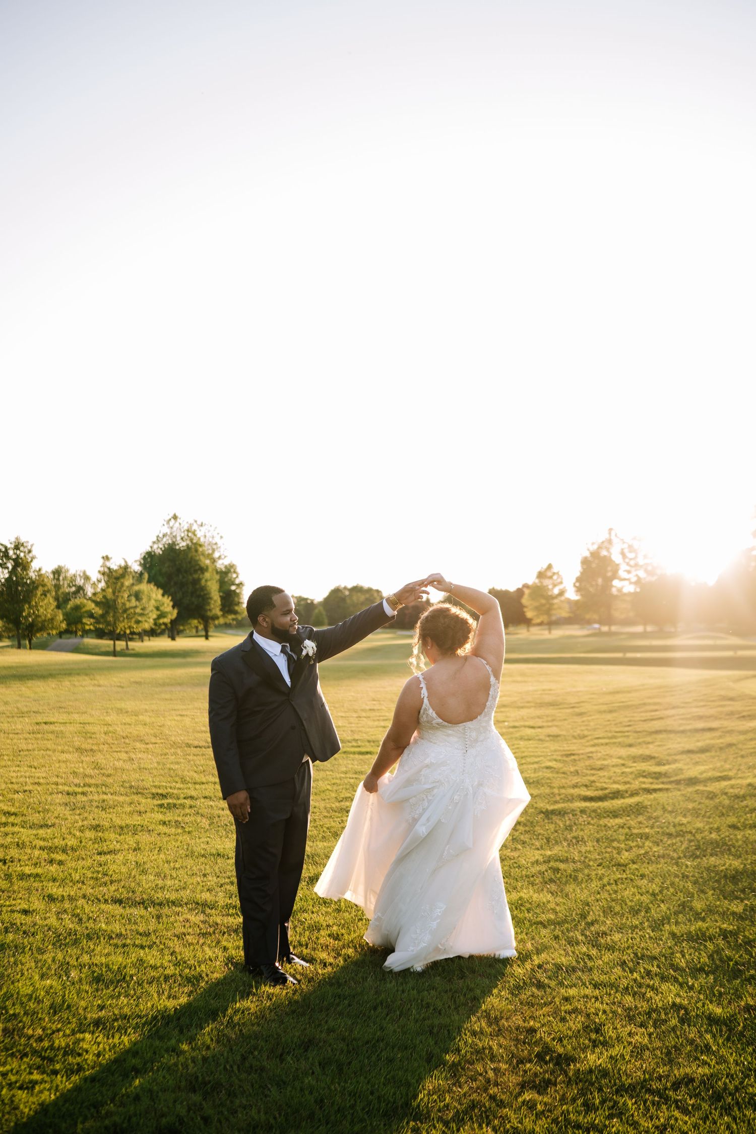 Newlyweds dancing together on grassy field during sunset.